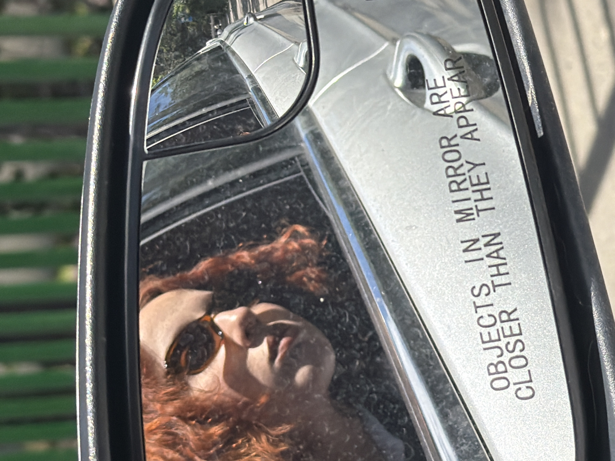 The image captures a side-view mirror reflecting a woman with curly red hair wearing sunglasses. The mirror's surface also shows a portion of the car's exterior. "Objects in mirror are closer than they appear" is imprinted on the mirror. In the blurred background, a green park bench is visible. The location is identified as Tbilisi, Georgia.