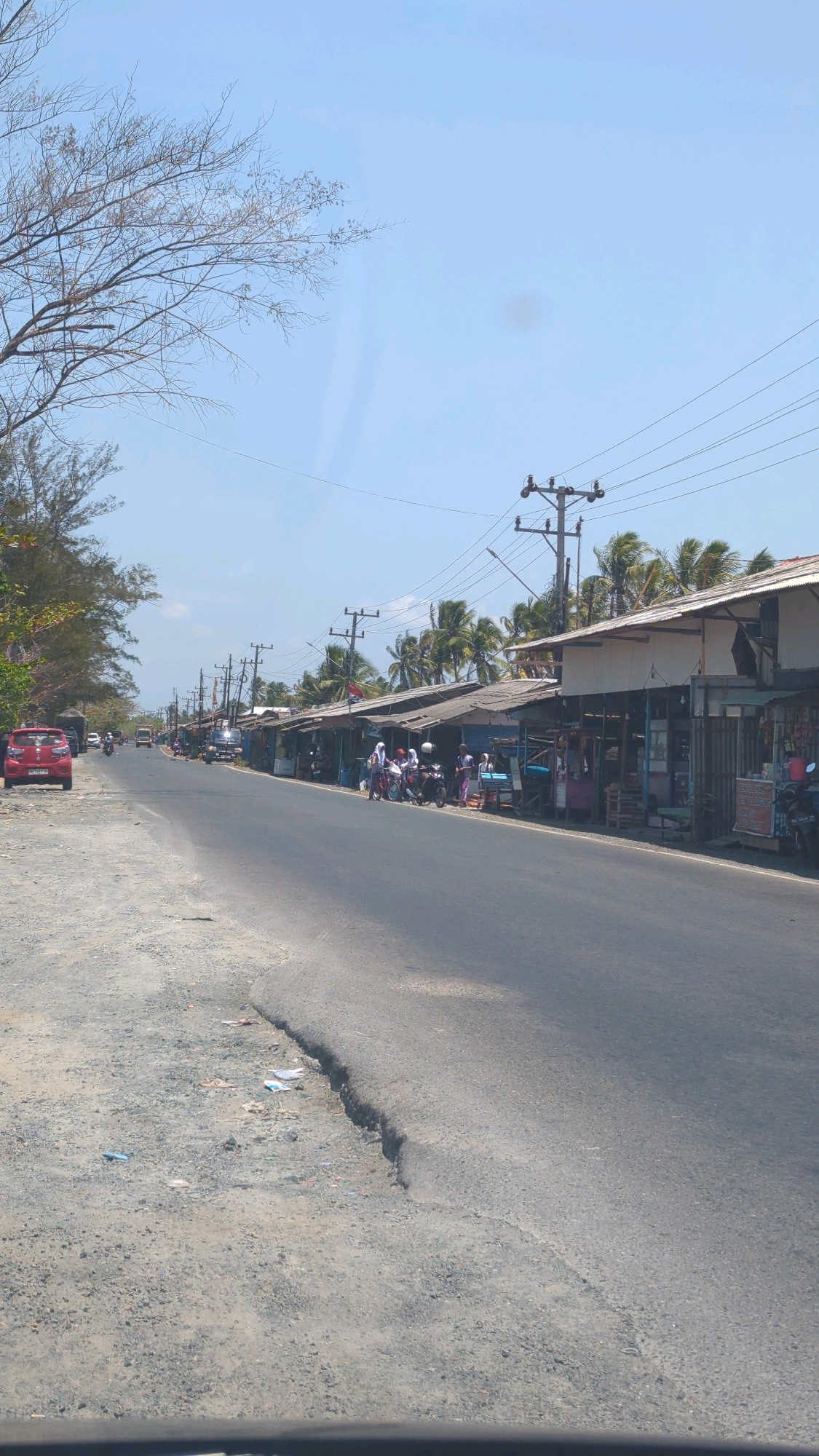The image depicts a street scene in a rural area, likely a village, in Beringin, Indonesia. A paved road runs through the center of the frame, with a gravel shoulder on the left and a row of small shops or stalls lining the right side. The shops have makeshift roofs and appear to sell various goods. Several people are visible, some standing near the shops and others on motorcycles. A red car is parked on the left side of the road. Palm trees and other vegetation are present, suggesting a tropical climate. Utility poles with overhead wires are visible along the road. The sky is clear and blue, indicating a sunny day. The overall impression is of a simple, functional roadside settlement.