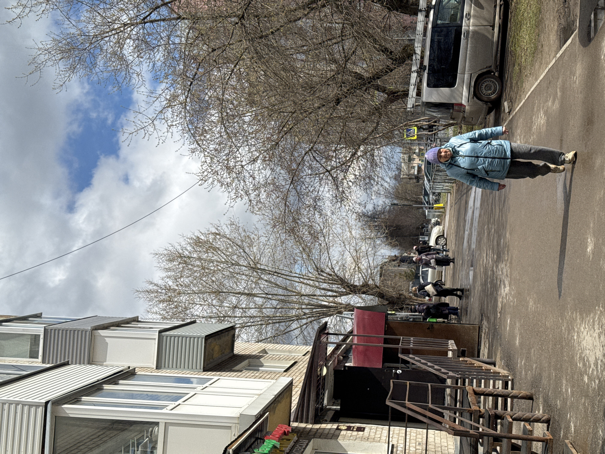 On a sunny day in Perm, Russia, a middle-aged woman wearing a blue jacket and purple hat walks on a sidewalk adjacent to a building. The building has multiple windows and an exterior staircase leading to a commercial entrance. People walk along the street further down, where cars are parked along the curb. A silver van with a ladder on its roof is parked closer to the camera, near a crosswalk. Bare trees line the street, with branches overhead against a backdrop of blue sky and white clouds.