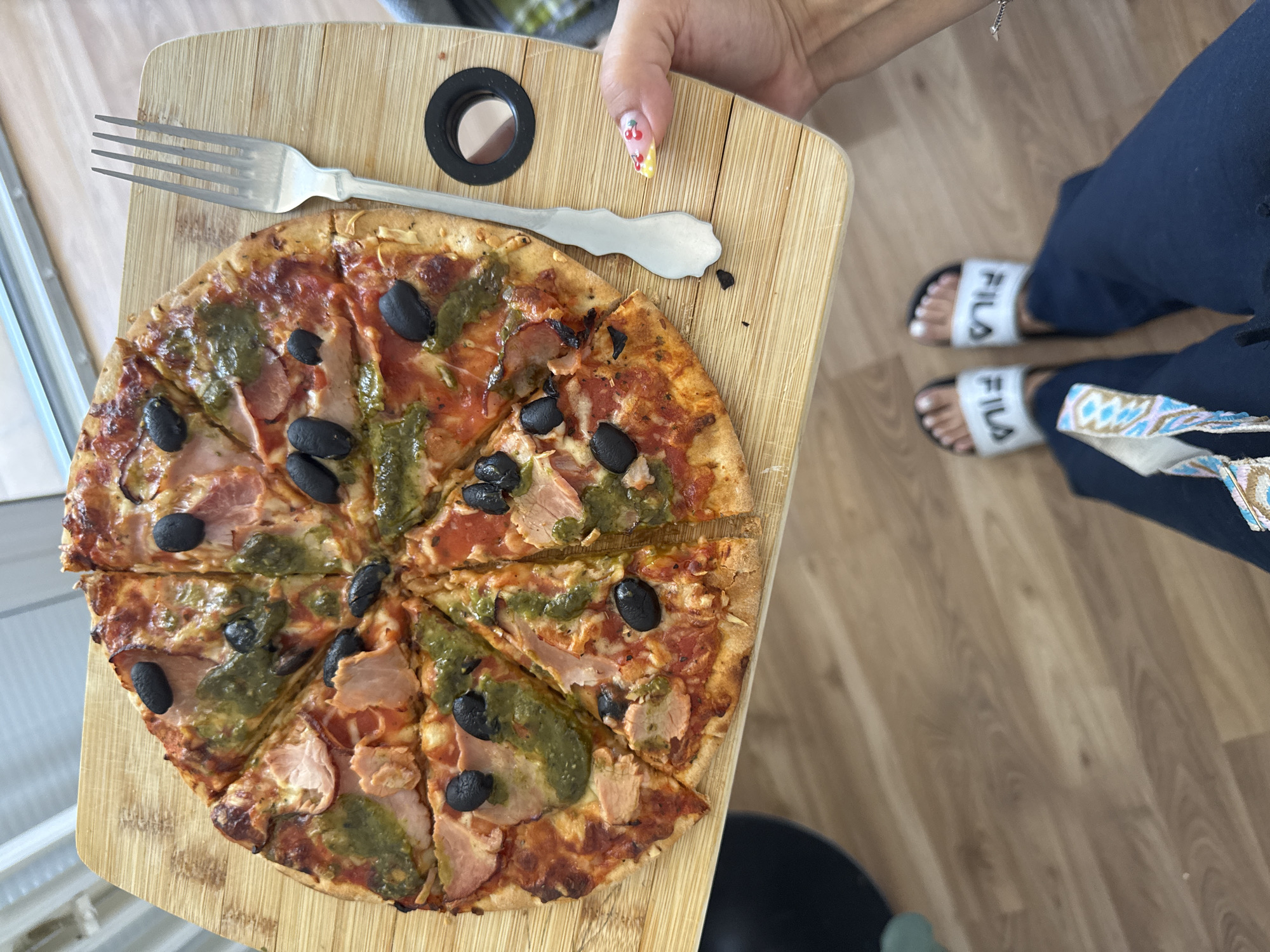 The image shows a close-up overhead view of a pizza on a wooden cutting board. The pizza is cut into eight slices and topped with tomato sauce, ham, black olives, and a green sauce, possibly pesto. A silver fork rests to the right of the pizza. A person's hand with decorated fingernails is visible holding the cutting board. Below the cutting board, the person's legs are visible, wearing dark blue pants and white Fila slides on their feet. The background consists of a light-colored wooden floor. There is no information in the image to suggest the location is Bruchköbel, Germany.