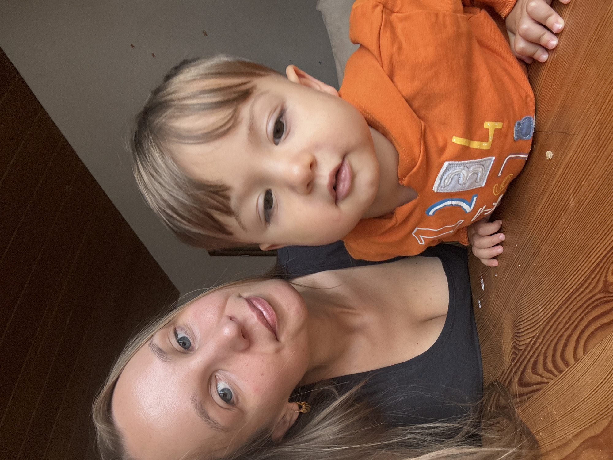 A woman and a toddler are on a wooden table. The woman, on the left, has blonde hair and is wearing a dark top. She is looking at the camera with a slight smile. The toddler, on the right, is wearing an orange hoodie with designs on it. The toddler is also looking towards the camera. The background shows a wall and a wooden ceiling. The image appears to be taken indoors, likely in a home in Überherrn, Germany.