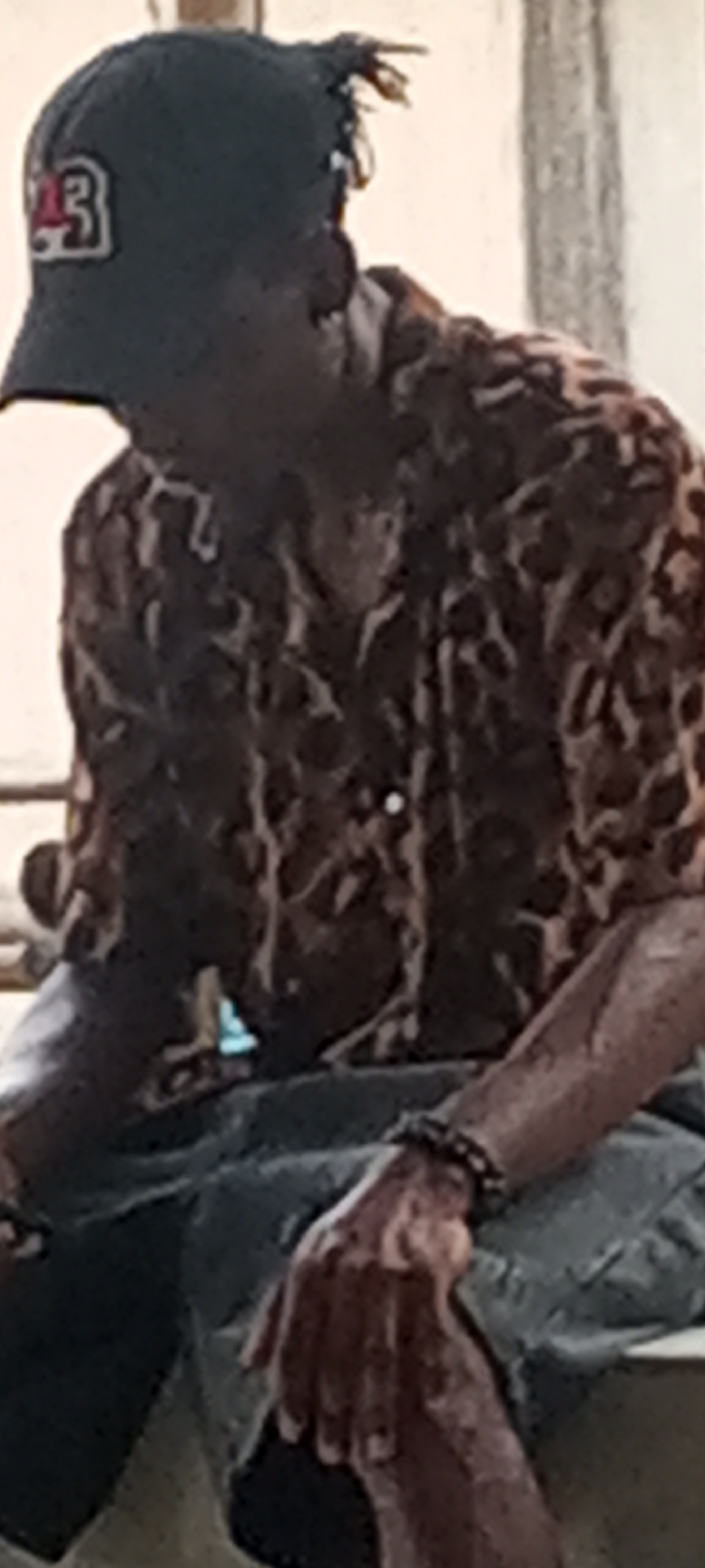 A person wearing a dark baseball cap with a number "23" on the front, a brown leopard print shirt, and grey shorts is seated. The location is Unknown, Unknown. A beaded bracelet is visible on the person's left wrist. The person's left hand rests on their lap. The individual's face is obscured by the hat.