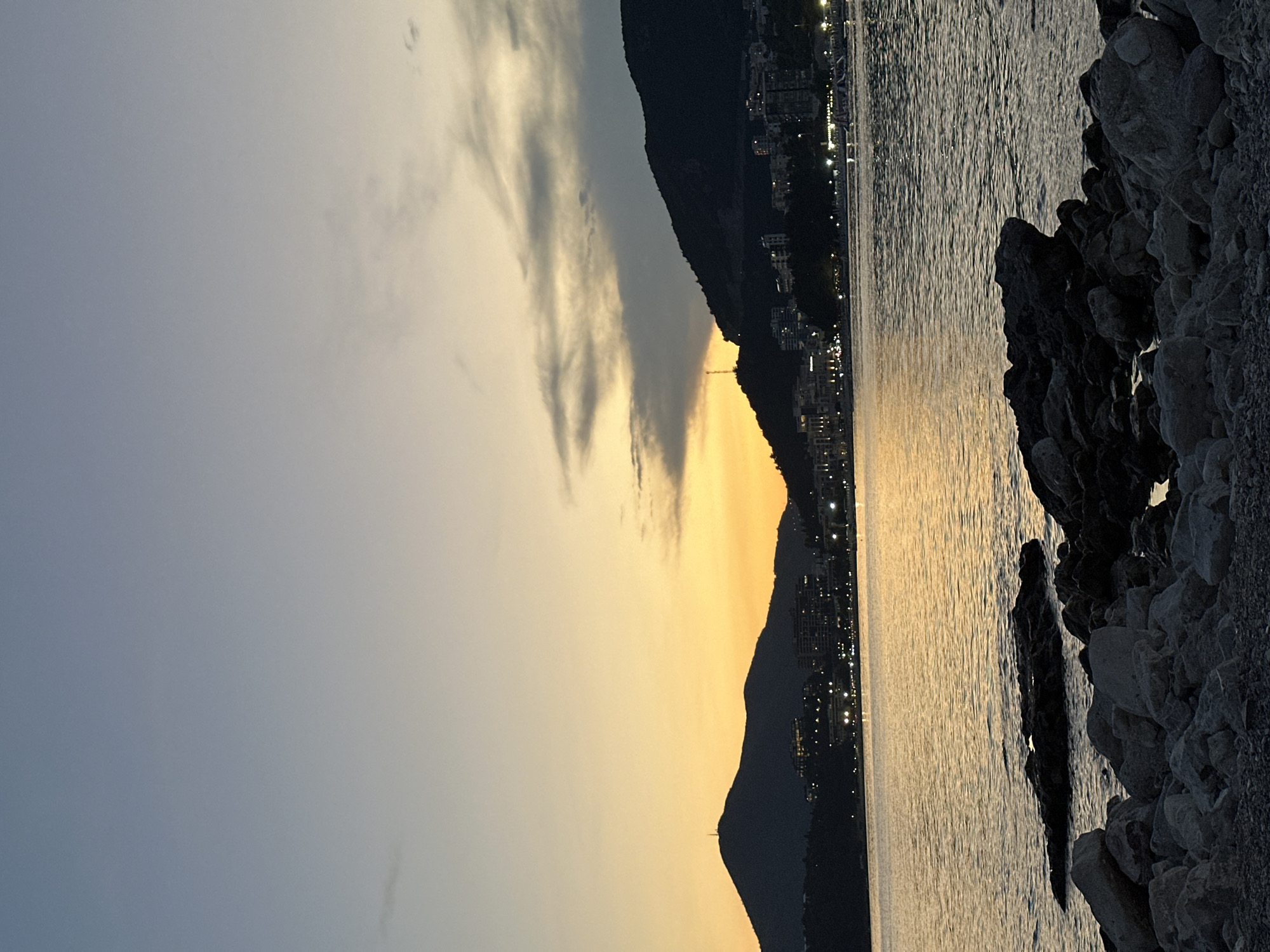 The image depicts a sunset over the Adriatic Sea in Rafailovići, Montenegro. The sky transitions from a pale blue at the top to a vibrant orange and yellow gradient near the horizon. Dark, silhouetted mountains rise from the coastline, with a city's lights twinkling along the shore. The foreground is a rocky beach with numerous stones of varying sizes, some submerged in the gently rippling water. The water reflects the warm hues of the sunset. The overall scene conveys a peaceful, twilight ambiance.