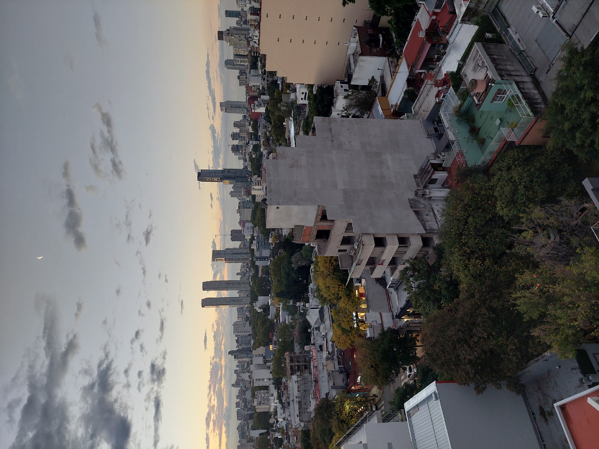 The image depicts an urban cityscape, likely taken from an elevated perspective. The view includes a dense collection of buildings ranging from low-rise residential structures to tall, modern skyscrapers. A variety of architectural styles are represented, from older buildings with tiled roofs to more contemporary concrete constructions. Trees and green spaces are interspersed among the buildings. The sky is visible in the background, showing clouds and a setting sun. A crescent moon is visible in the sky. The scene appears to be set in Buenos Aires, Argentina, based on the architectural styles and urban layout.