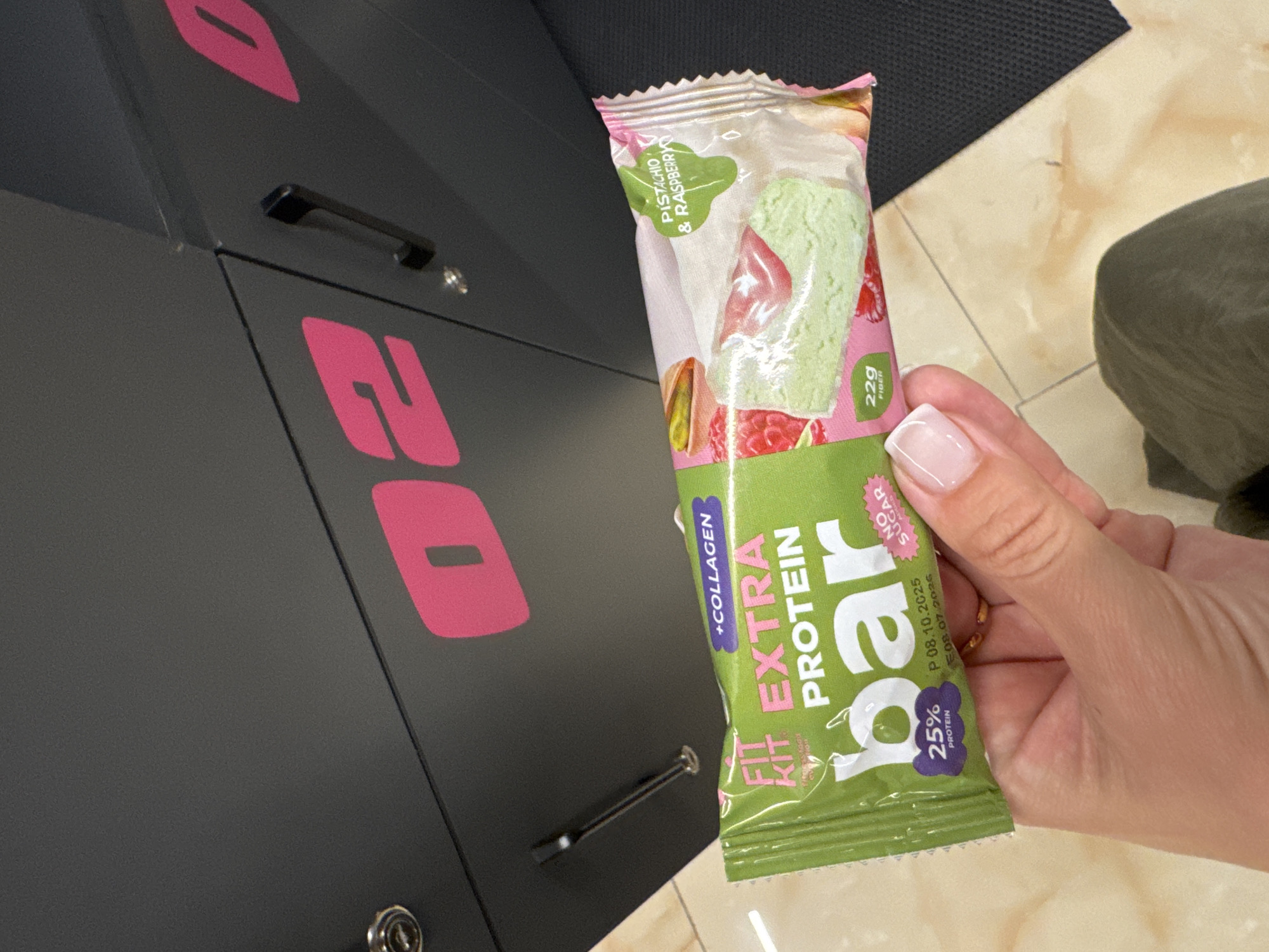 A woman's hand holds a "Fit Kit Extra Protein Bar" which is pistachio and raspberry flavored. The bar's packaging is predominantly green and features text indicating it contains collagen, 22g of fiber, 25% protein, and is "No Sugar." The background shows a portion of a dark gray locker with a pink "02" logo. The floor is tiled with a beige and cream pattern. The lighting suggests it is daytime. The image is cropped to focus on the protein bar and the hand holding it.