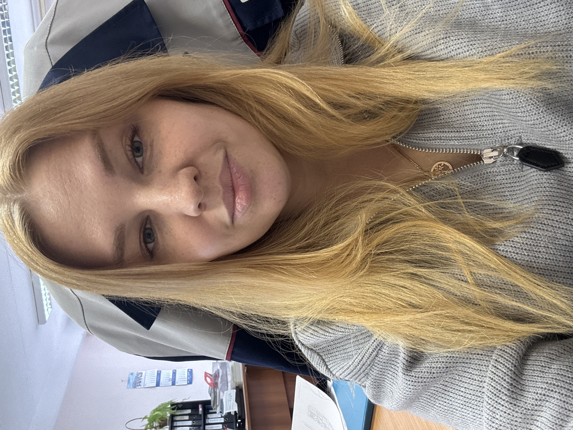 The image is a selfie of a fair-skinned woman with light blonde hair, wearing a gray zippered sweater and a gold necklace. She is indoors, likely in an office setting. The background includes a desk with various office supplies, a calendar on the wall, and a gray and blue office chair. There is no information in the image to indicate this photo was taken in Rusanovo, Russia.