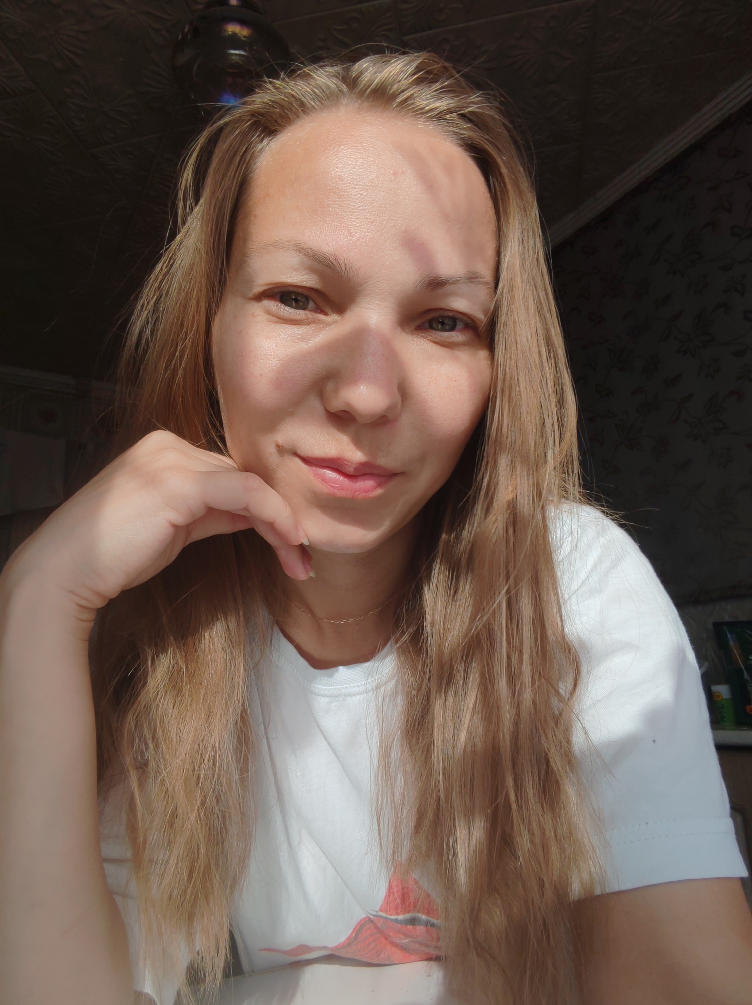A medium shot, portrait-style image shows a young woman with long, blonde hair, looking at the camera. She is wearing a white t-shirt. Her left hand is resting on her chin, and her right arm is down. Sunlight casts shadows across her face, creating a dappled effect. The background is dark and out of focus, with hints of wallpaper and a ceiling fixture visible. No specific location in the city of Чухлэм, Russia, can be identified from the image.