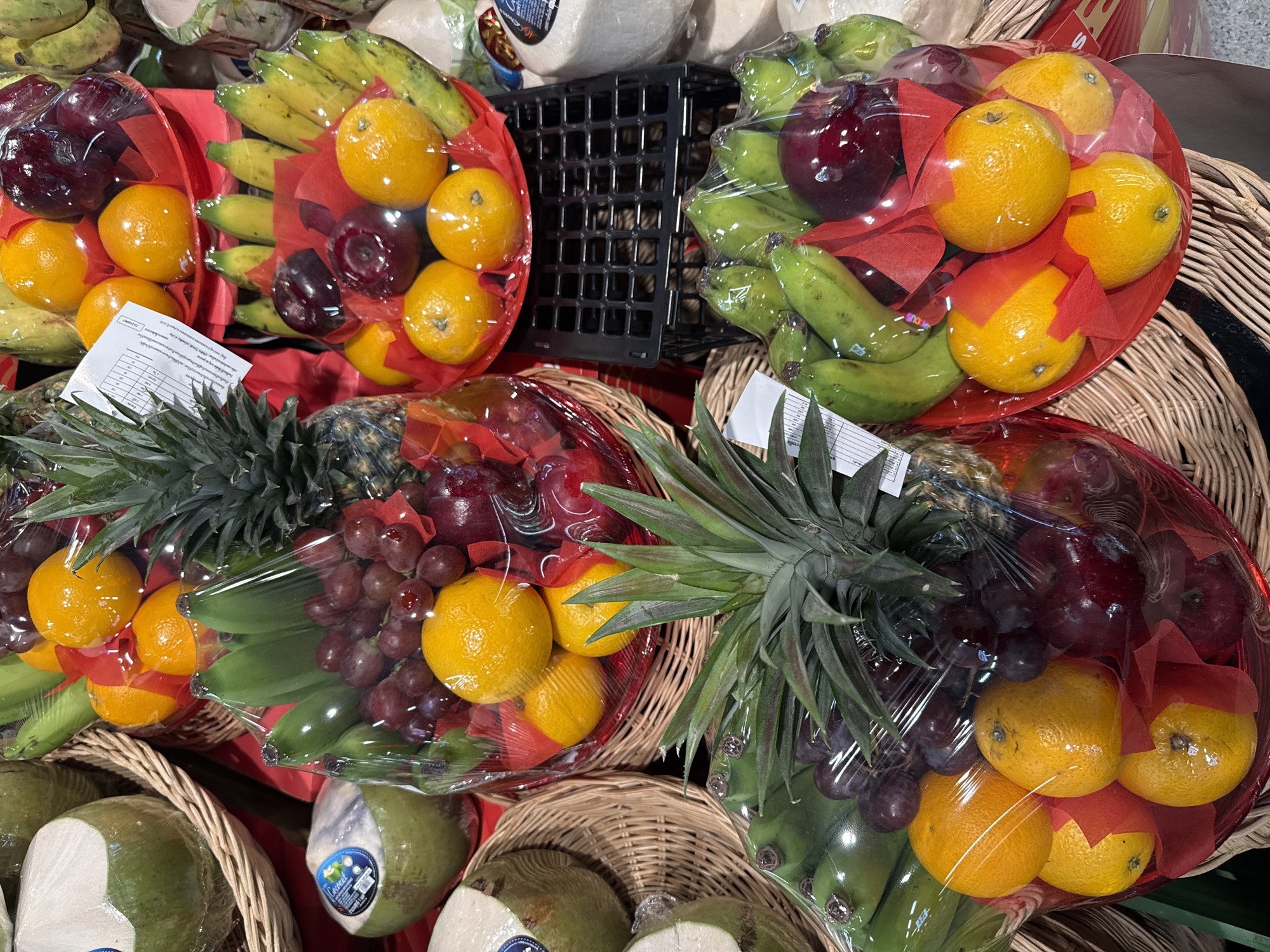 The image displays an assortment of fruit baskets, possibly in a market setting in Pattaya City, Thailand. The fruits are predominantly red and yellow, with some green and purple varieties. Several baskets are visible, containing items like oranges, apples, grapes, pineapples, and bananas. The fruits are wrapped in clear plastic and adorned with red decorative elements. The scene appears to be an outdoor or semi-outdoor market, judging by the baskets and the variety of produce. The image is taken from a slightly elevated angle, looking down at the fruit displays. No people are visible in the image. The overall impression is one of fresh produce arranged for sale.