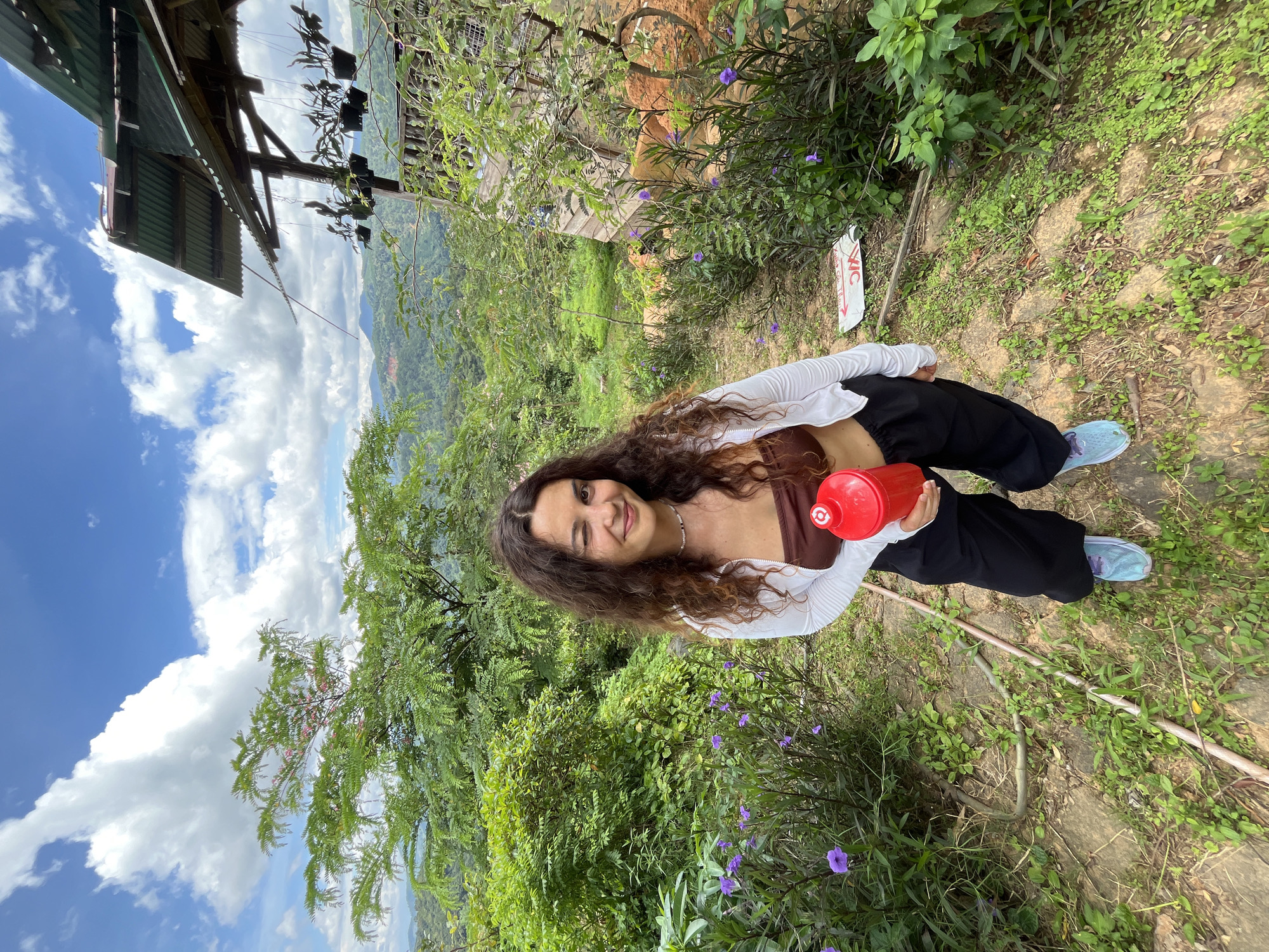 A woman with long, curly brown hair is standing outdoors, winking at the camera and holding a red water bottle. She is wearing a brown strapless top, a white cardigan, black pants, and light blue sneakers. The background shows a lush, green landscape with rolling hills and mountains, indicative of the Sơn Điền area in Vietnam. A rustic, open-air structure with a corrugated metal roof and hanging plants is visible to the right. The ground is covered with dirt, gravel, and patches of green vegetation, including small purple flowers. The sky is blue with scattered white clouds.