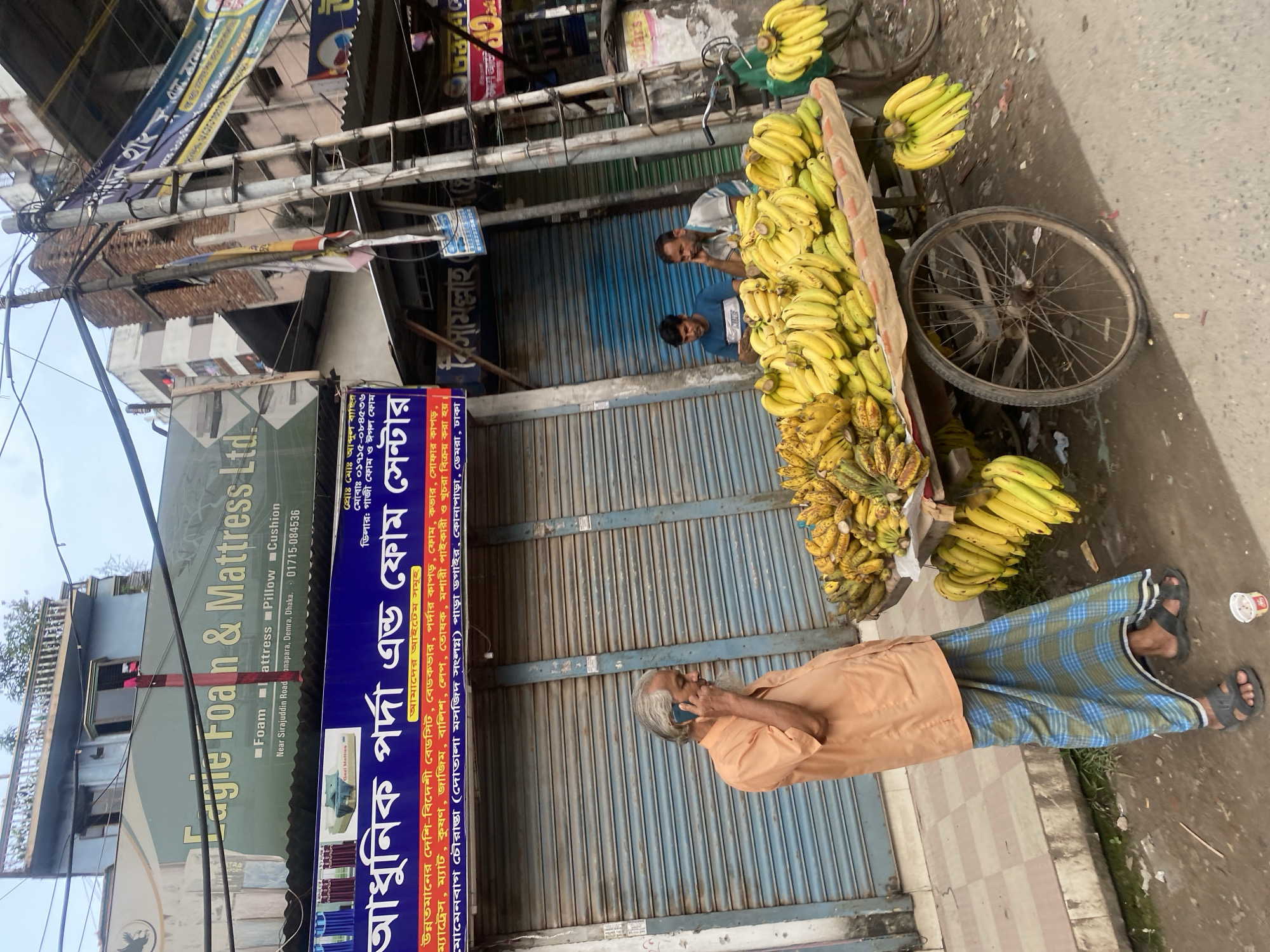 In Dhaka, Bangladesh, an outdoor scene unfolds with a man in an orange shirt and a lungi talking on a mobile phone, standing next to a cart overflowing with bananas. The cart, also laden with bananas hanging from its sides, is parked on a street lined with shops. In the background, two other men are seated near the banana cart, one in a blue t-shirt and the other in a striped shirt. Overhead, power lines and electrical poles crisscross the sky, and a tall building looms above the storefronts. The overall impression is a bustling street market in the city.