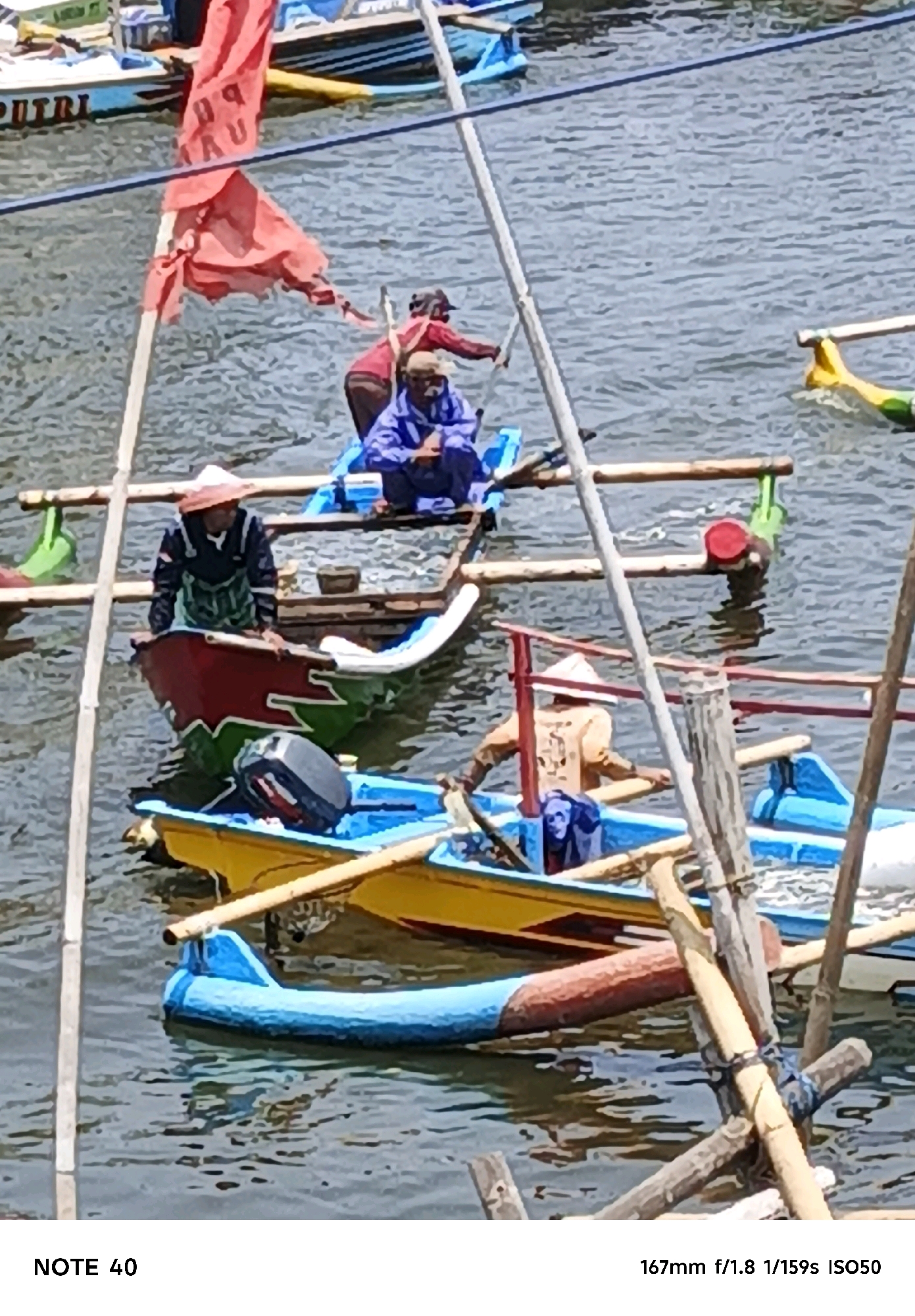 In the waters of Kelurahan Tegalkamulyan, Cilacap, Indonesia, several wooden boats are docked or in motion. One boat in the foreground, painted yellow with a blue inflatable section, has a person wearing a tan shirt and a conical hat paddling. Behind this, another boat, painted red and green with white accents, has a person in a dark shirt and patterned skirt standing. An outboard motor is visible on this boat. Further back, a blue boat is occupied by a person in a blue jacket. In the upper left, a boat with the word "PUTRI" is visible, with a red flag attached to a pole. The water is calm, with light rippling, and the scene appears to be a sunny day.