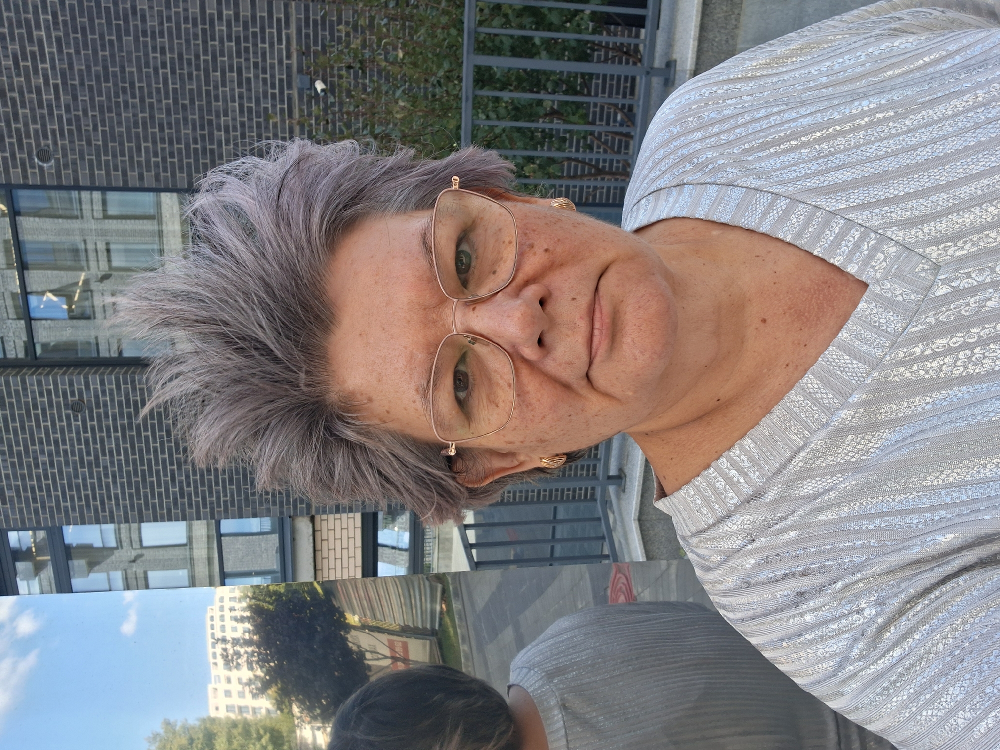 A middle-aged woman with short, spiky gray hair and glasses poses for a selfie. She is wearing a silver, V-neck top with a textured pattern. The background shows a modern brick building, a metal fence, and some greenery. A large, reflective surface to the left of the woman shows a reflection of her back and shoulder, as well as buildings and trees in the distance. The environment appears to be an urban setting, possibly a residential or commercial area. The image was taken outdoors, with a cloudy sky visible in the reflection. No specific location details are discernible from the image itself, and therefore, the statement about it being in Novosibirsk, Russia cannot be confirmed from the visual information provided.