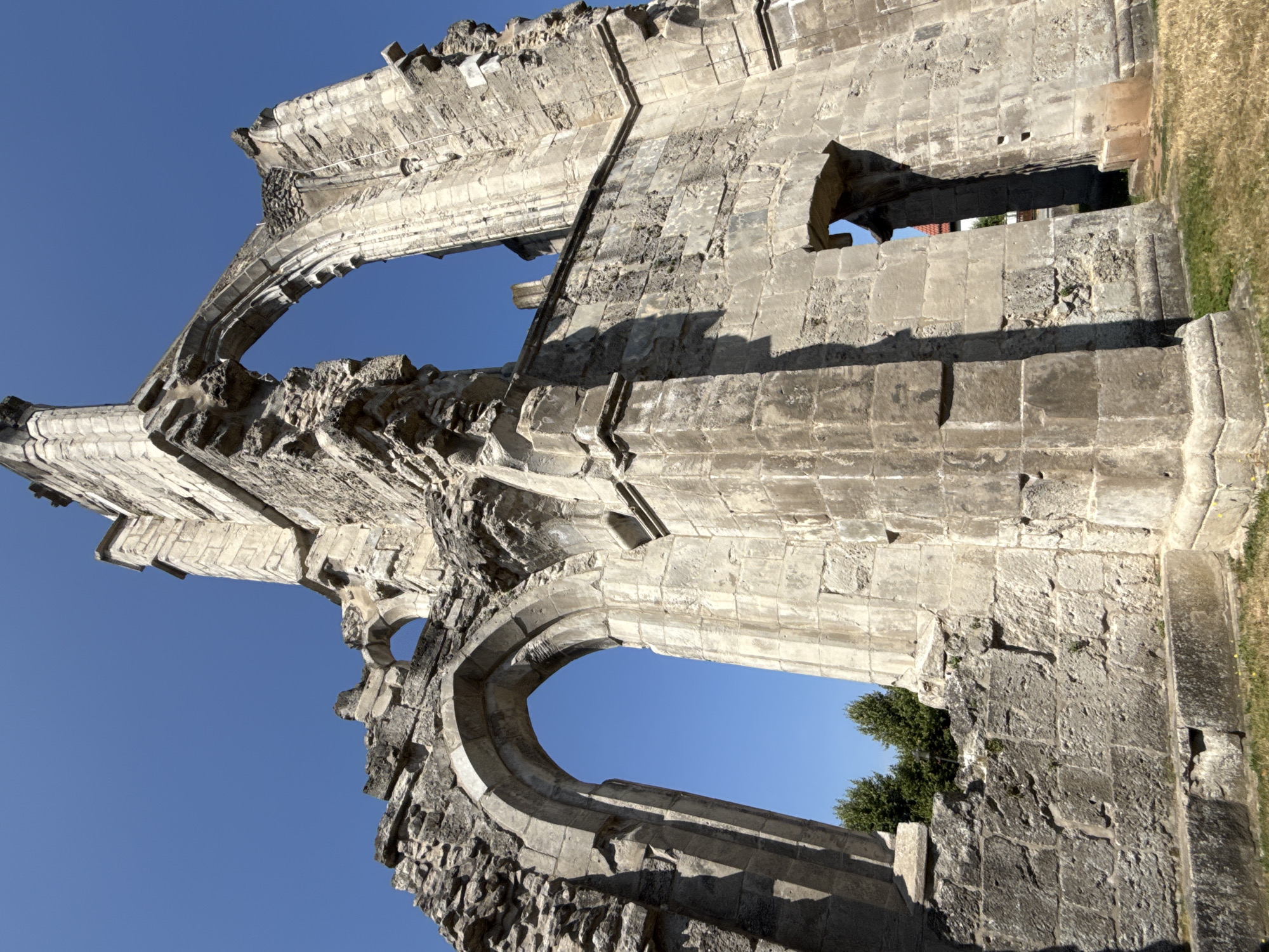 The image shows the exterior of the ruins of the former Walkenried Abbey in Walkenried, Germany. The ruins are constructed of large, weathered stone blocks and feature imposing arched openings that reveal a clear blue sky. The architecture suggests a historical religious structure, likely Gothic in style, with evidence of significant decay and partial collapse. Sunlight casts strong shadows across the stonework, highlighting the textures and irregularities of the ancient building. The ground in front of the ruins is covered with dry grass and a few small yellow flowers. In the background, the tops of green trees are visible beyond the walls, and a hint of a red-tiled roof can be seen through one of the arched openings. There are no people or animals present in the scene.
