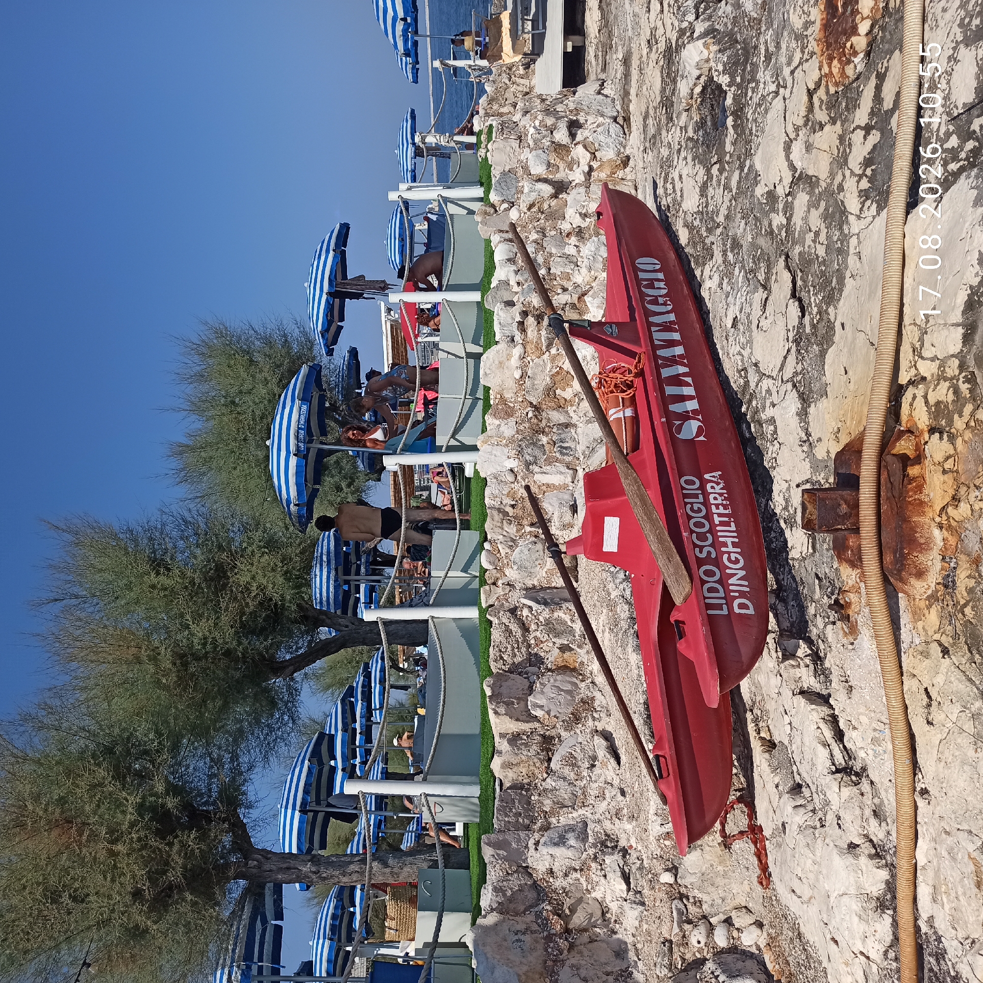 A bright, sunny day at Lido Scoglio D'Inghilterra, Molfetta, Italy, is depicted. In the foreground, a red lifeboat with oars rests on rocky terrain. The boat has "LIDO SCOGLIO D'INGHILTERRA SALVATAGGIO" written on its side. Behind the lifeboat, a stone wall separates the rocky area from a grassy area with beach umbrellas and loungers. Several people are seen relaxing under the umbrellas, with some individuals interacting. The sea is visible in the background. Trees with sparse foliage are scattered throughout the scene, providing shade. The timestamp "17.08.2026 10:55" is present in the bottom right corner.