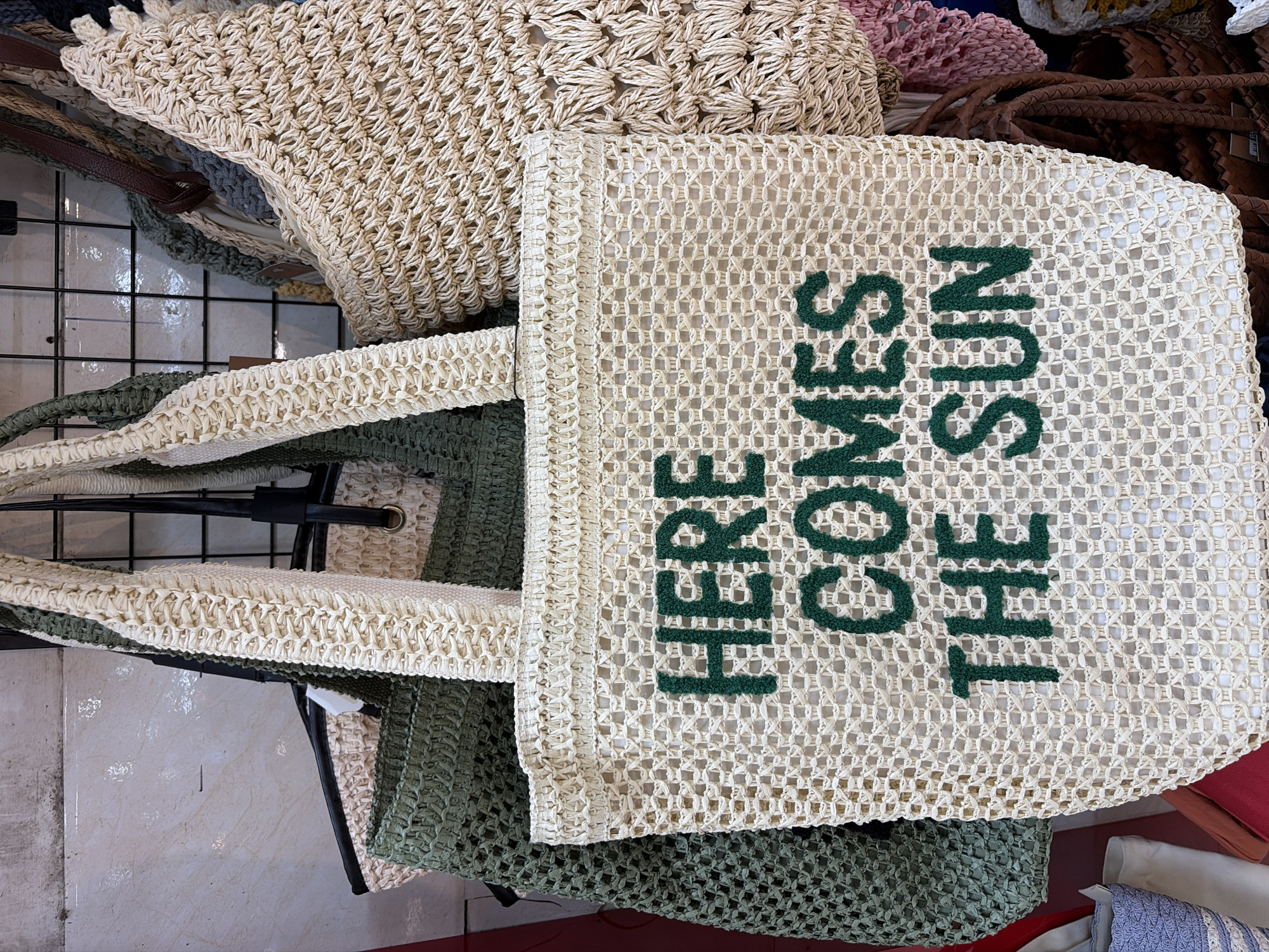 A close-up, eye-level shot shows several woven tote bags hanging from a wire rack. The bag in the foreground is off-white with green embroidered lettering that reads "HERE COMES THE SUN." To the left of this bag is a green woven tote bag, and behind it is another off-white woven tote bag with a textured pattern. Other bags, in various shades of beige, pink, and brown, are visible in the background. The scene appears to be a market stall. The image was taken in Da Nang, Vietnam.