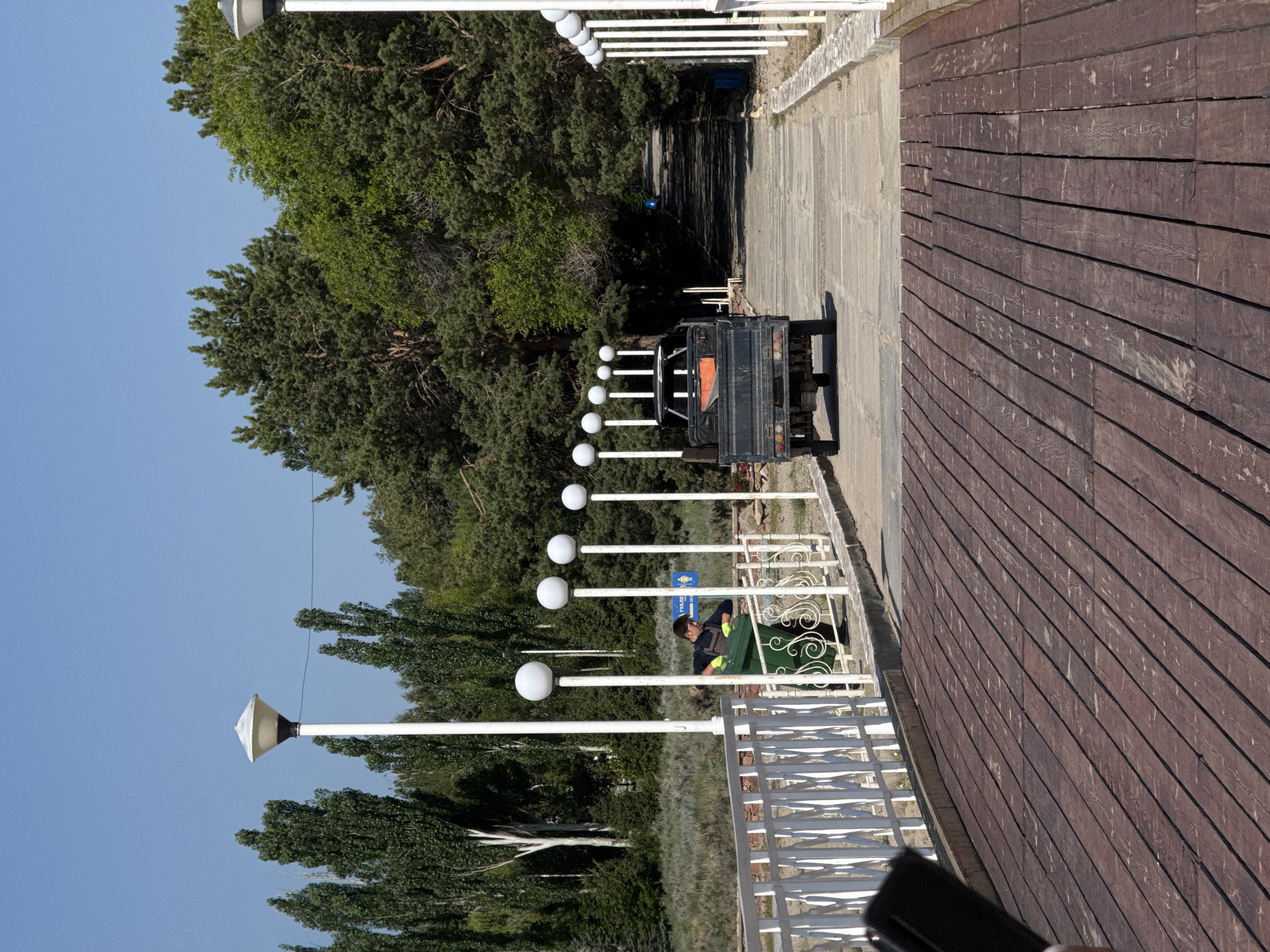 In Koshkol', Kyrgyzstan, a scene unfolds on a paved pathway lined with white railings and spherical lamps. A man, wearing a dark vest over a blue shirt and yellow gloves, is engaged in emptying a large green garbage bin into a dark-colored, three-wheeled vehicle. The vehicle is parked on the pathway, facing away from the viewer. The pathway itself is surrounded by lush green trees and a clear blue sky. The foreground is dominated by a wooden deck. In the background, a blue sign with white text and an icon is visible.