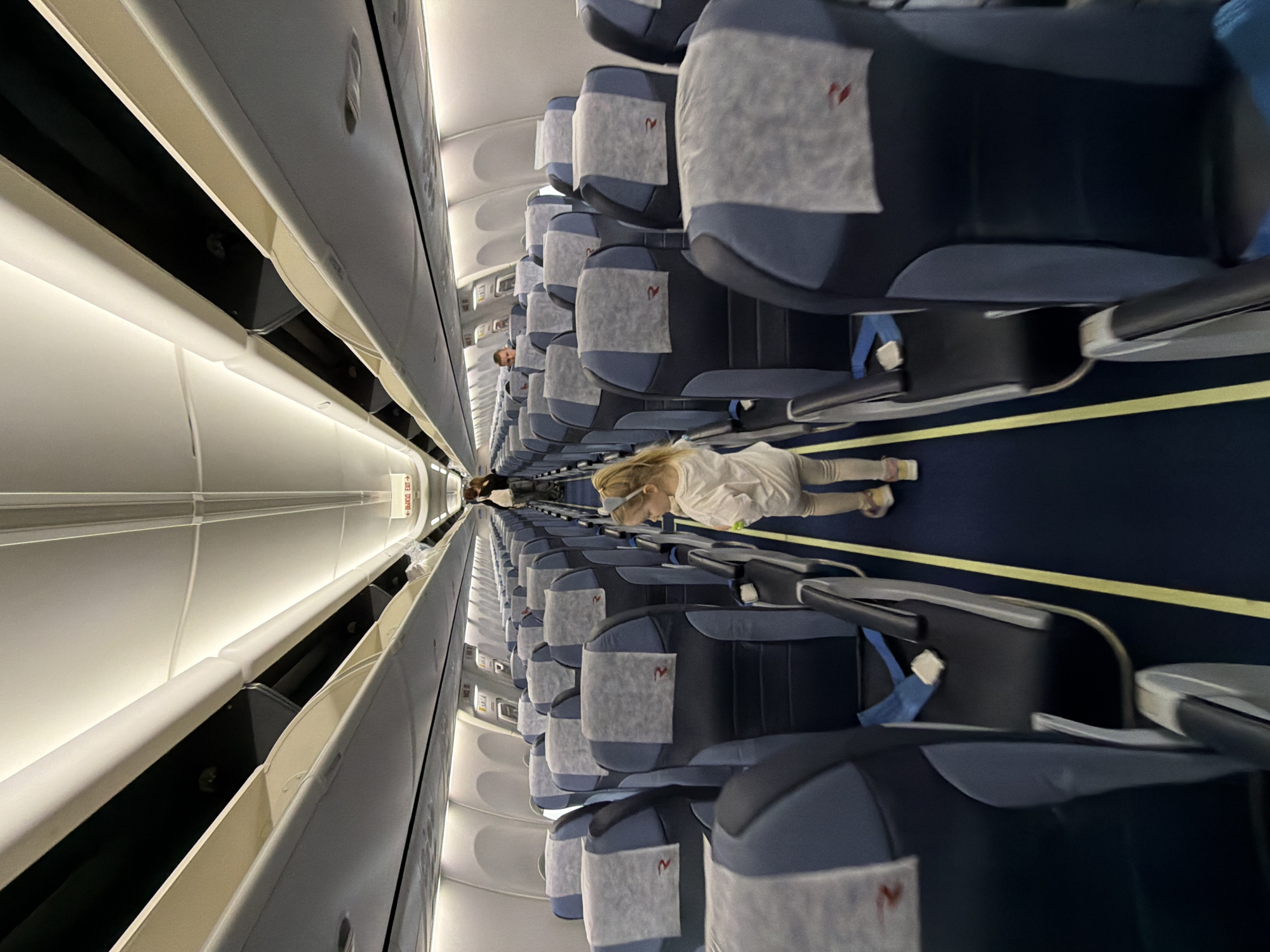 An interior shot of an airplane, likely a commercial airliner. The photo is taken from the aisle, looking toward the front of the plane. Rows of blue and gray seats are visible, with a light gray headrest cover on each seat. A young person with blonde hair is lying in the aisle. Some passengers are walking around in the distance. The plane's overhead compartments and interior lighting are visible. There is no evidence the photo was taken in Khimki, Russia.