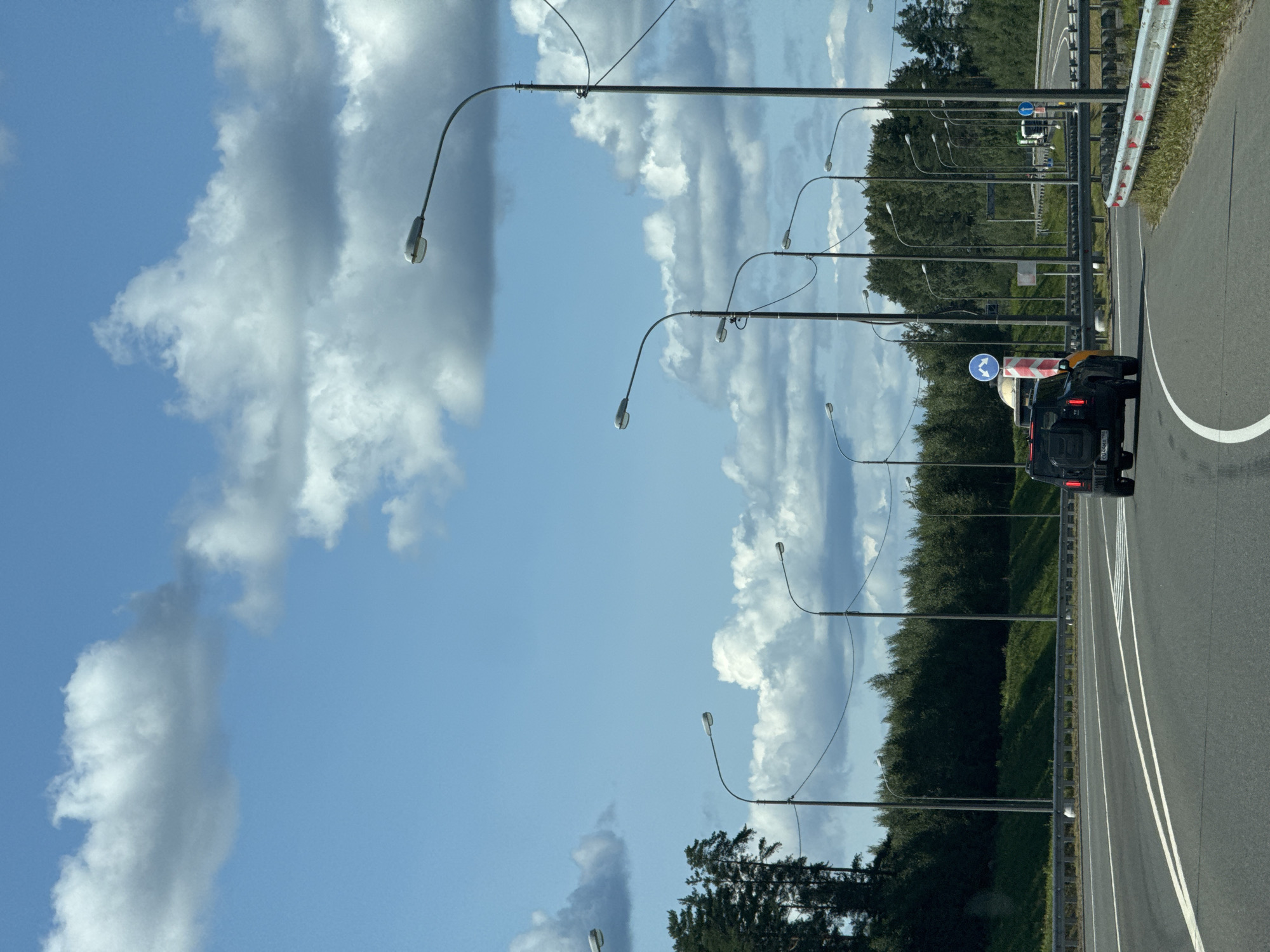 The image captures a view from a vehicle traveling on a highway with multiple lanes, likely in the city of Vaskelovo, Russia. The road is paved, with clear white lane markings. On the right side of the road, a metal guardrail with red and white reflective markers separates the highway from the grassy shoulder and bordering vegetation. On the left side, a dense line of trees forms a backdrop to the highway.
The sky is a vibrant blue with scattered white, fluffy clouds. Numerous streetlights on tall poles are visible along the highway, lining the road at regular intervals. The furthest streetlights appear to be part of a larger cluster indicating an intersection or exit.
In the foreground, a black SUV is driving away from the viewer in the right lane. The SUV has its taillights illuminated. Further ahead on the highway, other vehicles are visible, including what appears to be a truck and several cars. A traffic sign with blue arrows indicating a diversion or lane change is also visible on the right. The overall scene depicts a clear, sunny day on a well-maintained highway infrastructure.