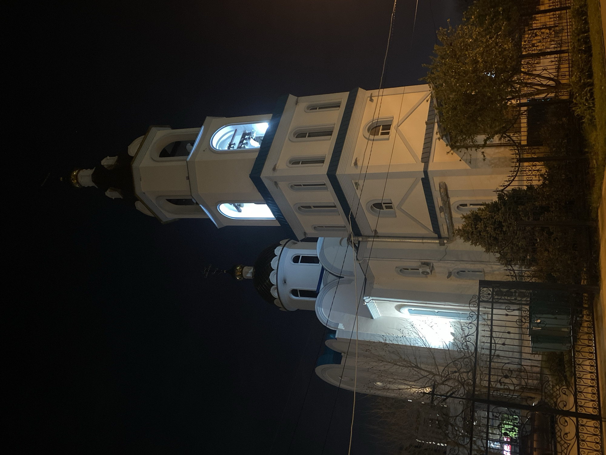 At night, an exterior view of a brightly illuminated white church with blue accents and a black dome is shown behind a black metal fence. The church is on the right of the frame, while the sky is black. Trees are on either side of the fence.