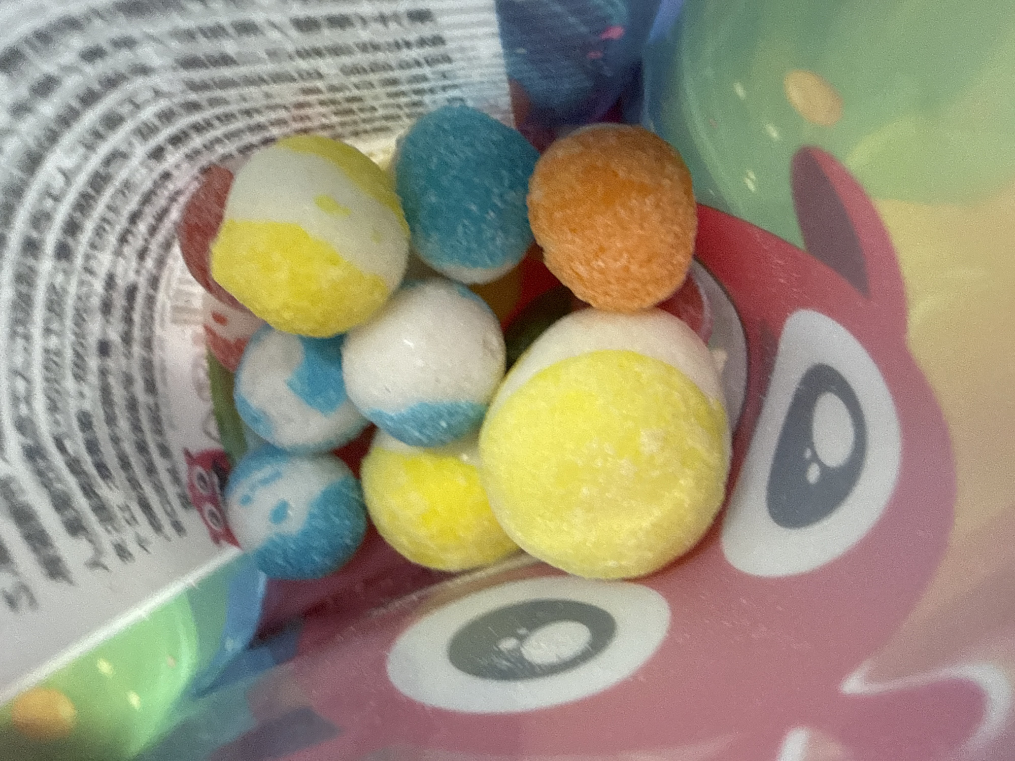The image displays a close-up view of several brightly colored, round candies scattered inside a pink bag with a cartoon character print. The candies are a mix of white, yellow, blue, and orange, some with multiple colors. The bag appears to be made of a plastic material and has a glossy finish. In the background, there is a blurred view of text on a white surface and a patterned interior of the bag. The scene does not contain any information or context related to Yakovlevskoye, Russia.