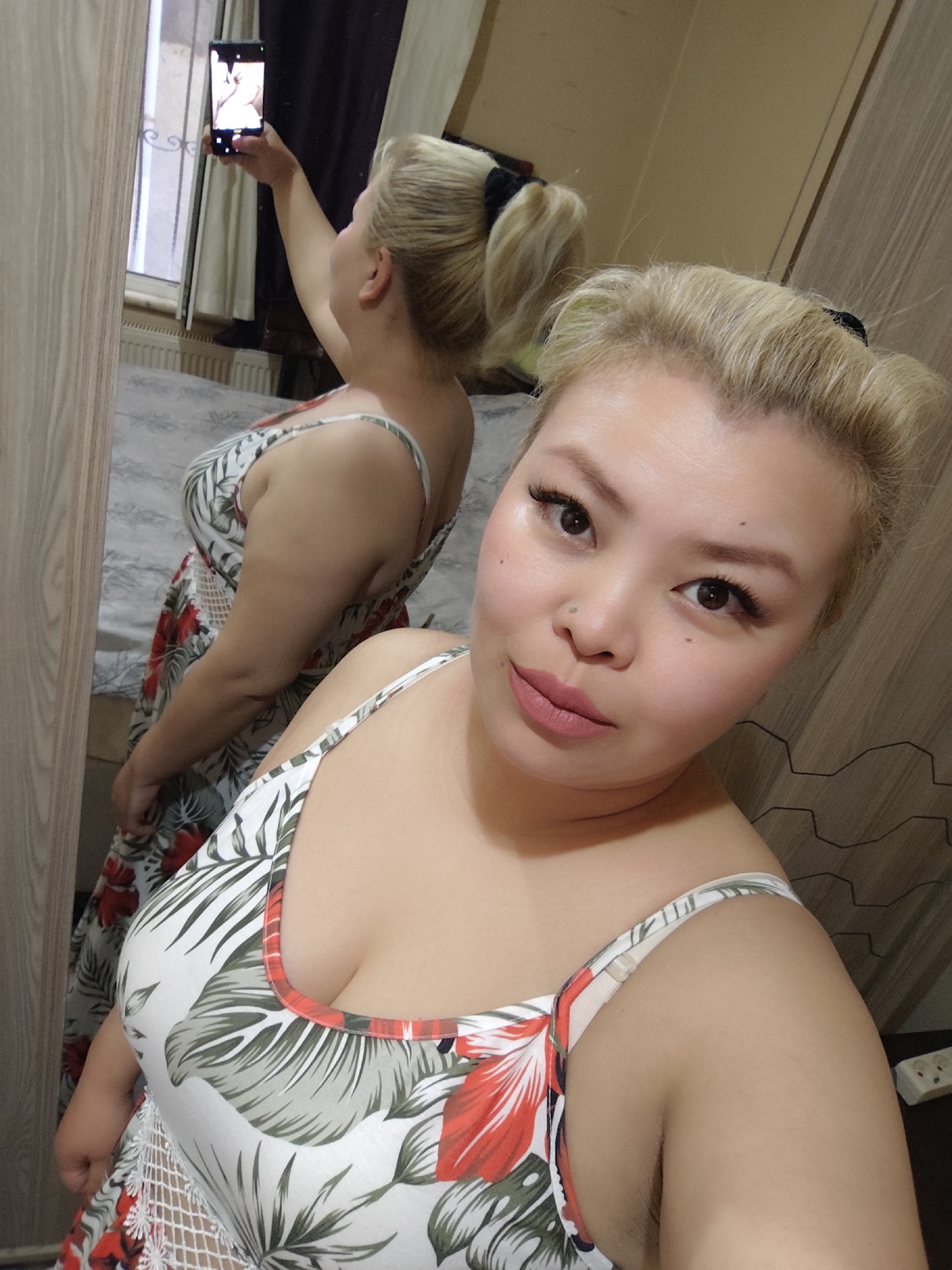 A woman is taking a selfie in front of a mirror. She has blonde hair pulled back in a ponytail with some hair styled upwards. She is wearing a floral print dress with white straps. In the mirror, the reflection shows her holding a cell phone to take the picture. Behind her in the reflection, a bed with a patterned bedspread is visible. The background includes a wooden wardrobe and a window with a radiator beneath it. The location appears to be indoors. The city of Sarıyer, Turkey cannot be determined from the image.