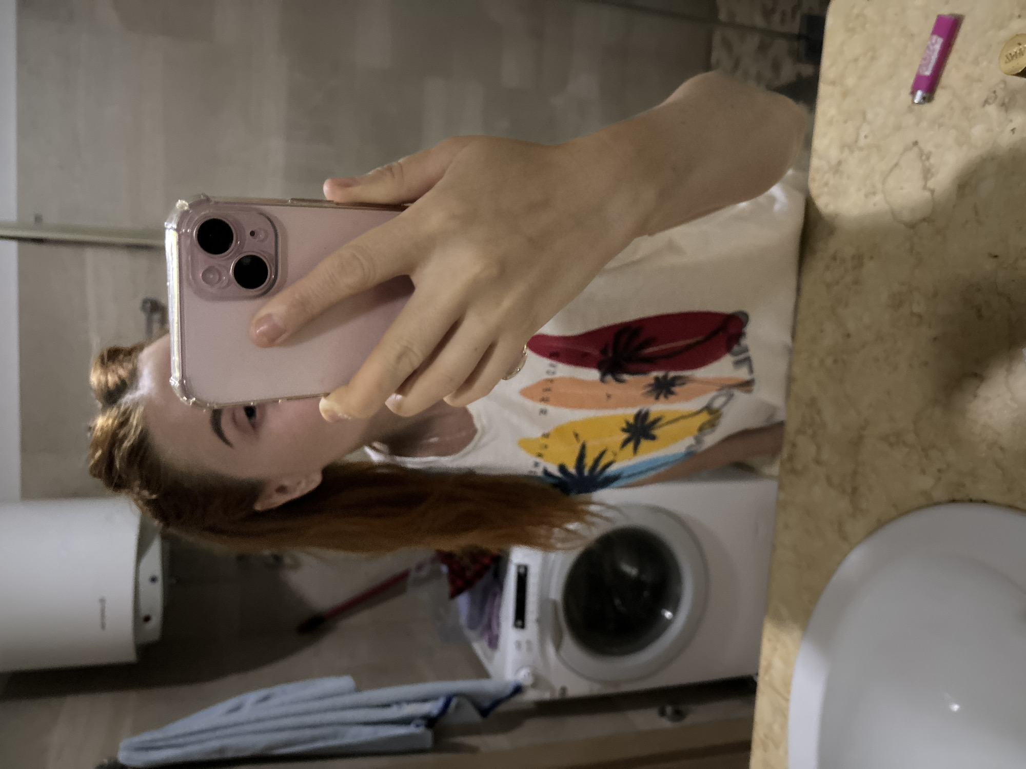 A woman with reddish-brown hair is taking a selfie with a pink iPhone in a bathroom. She is wearing a white T-shirt with a surfboard design. In the background, there is a washing machine, a white water heater, a sink, and a countertop with a pink lighter and a coin on it. The bathroom walls are gray. The location is likely a private residence. The city, Budva, Montenegro, is not discernible from this image.