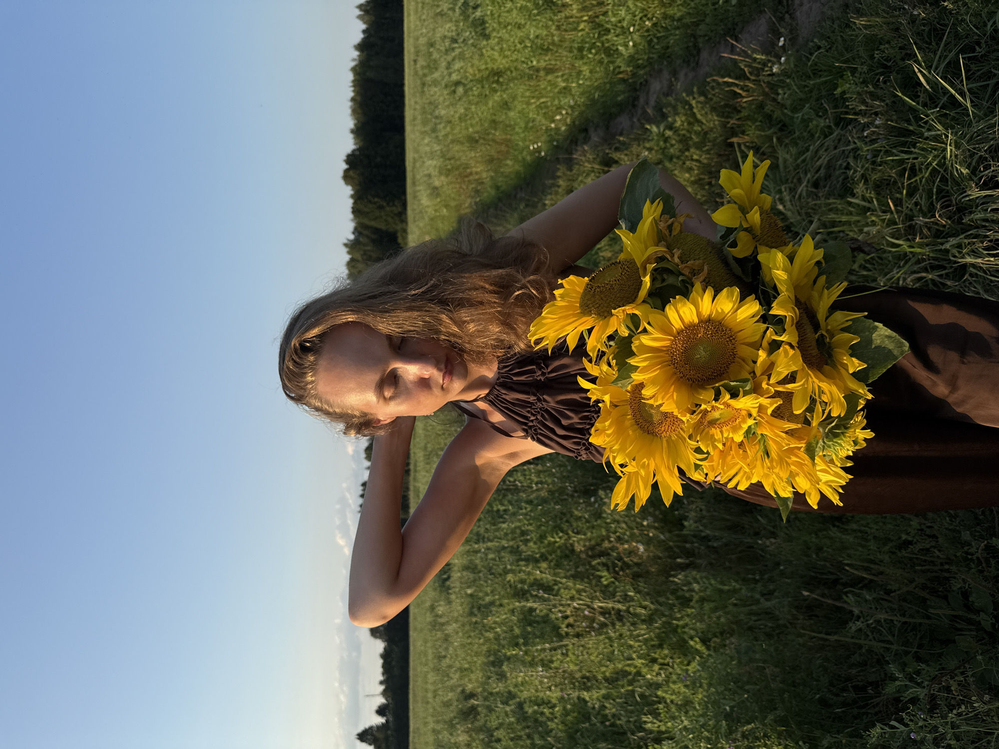 A young woman with long blonde hair is pictured outdoors, holding a bouquet of sunflowers. She is wearing a dark brown dress with a ruched neckline. Her eyes are closed, and her head is tilted back slightly, with her left arm raised behind her head, suggesting a relaxed or pensive mood. The sunflowers are bright yellow and fill the right side of the frame, obscuring part of her body.
The setting is a grassy field under a clear blue sky. In the background, a line of trees is visible along the horizon, and a dirt path can be seen to the left of the woman. The lighting suggests it is late afternoon or early evening, with the sun casting a warm glow on the scene. The overall atmosphere is serene and natural. The image appears to have been taken in Aksaitovo, Russia. There is no visible text in the media file.