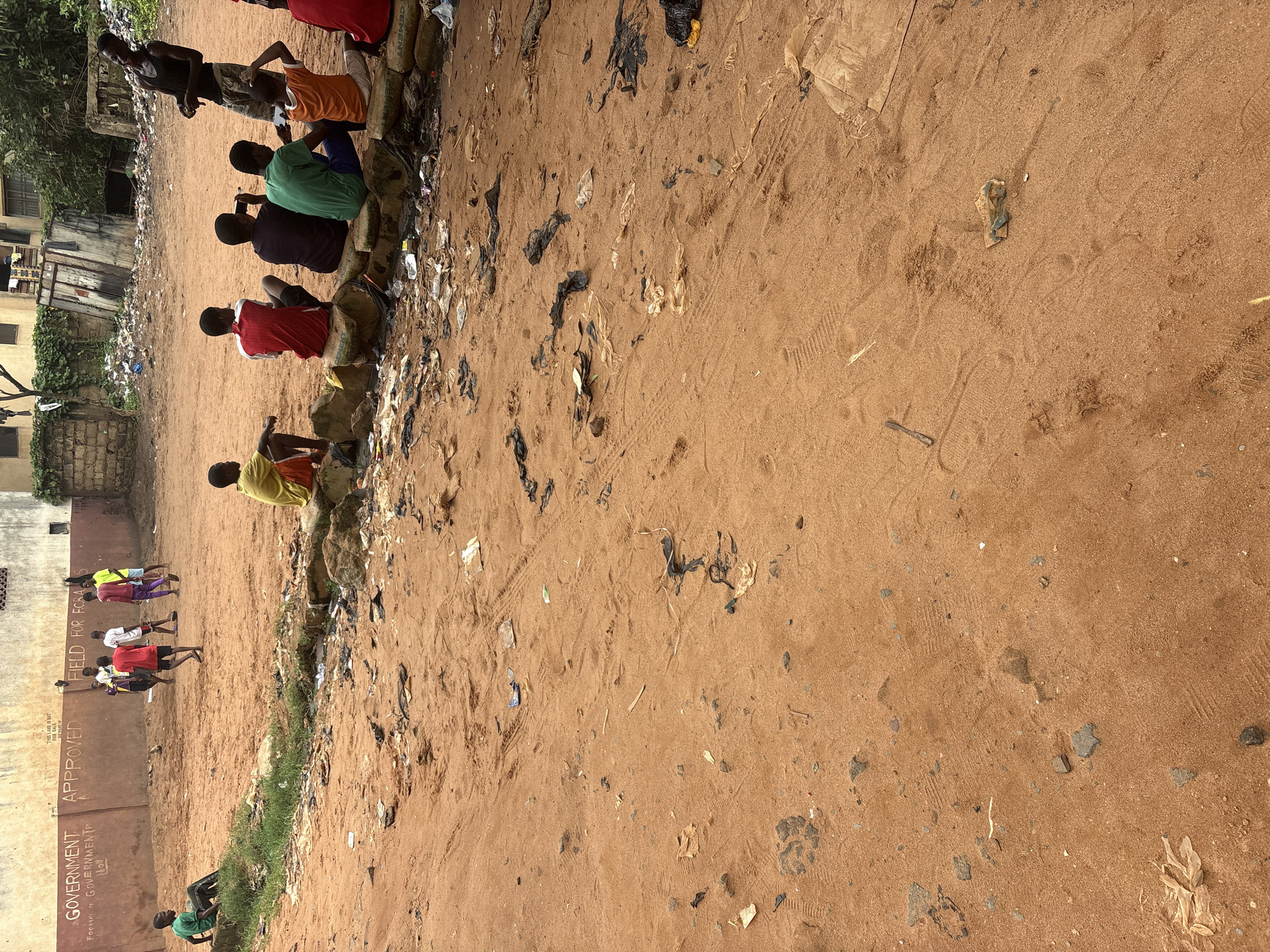 The image depicts a sandy area in Onitsha, Nigeria, characterized by a considerable amount of scattered litter and debris. A group of young males are present in the scene. Several are seated on a low wall constructed from cement bags, while others are standing and interacting in the sandy field. In the background, a wall with graffiti is visible, with text that includes "GOVERNMENT APPROVED FIELD FOR FCBA". The environment appears to be an unkempt outdoor space, possibly a makeshift sports field or recreational area, with elements of urban decay evident in the surrounding environment and the presence of waste.
