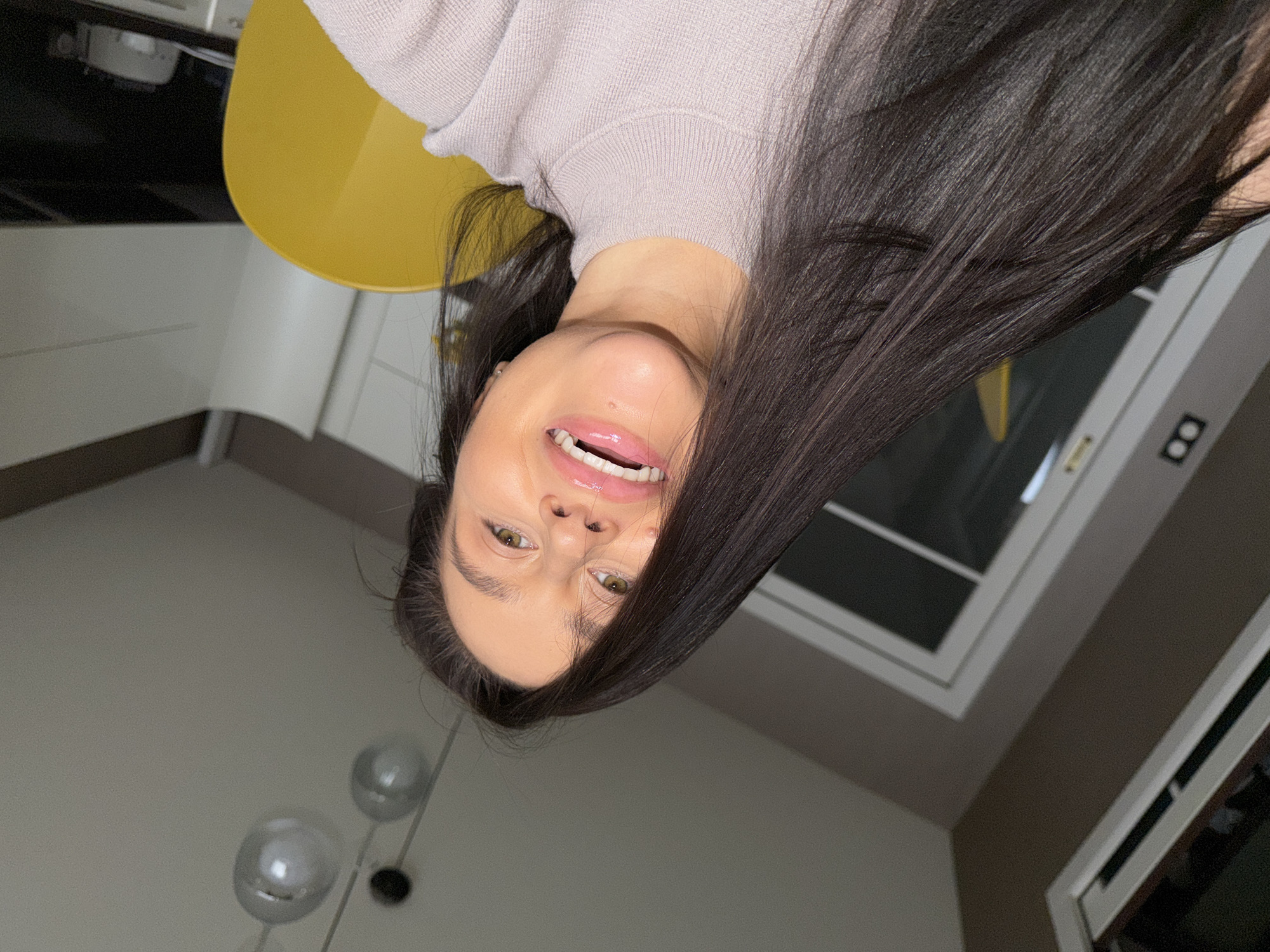 An indoor portrait features a young woman, likely in her 20s or early 30s, looking upwards into the camera with a cheerful, broad smile that reveals her teeth and glossed lips. She has long, straight, dark hair that cascades around her shoulders, partially obscuring the left side of her face. Her eyes are light-colored, possibly hazel or green. She is wearing a light-colored, possibly light purple or grey, ribbed knit sweater.
The setting is a modern home interior, suggesting a kitchen or dining area in Khimki, Russia. Behind her, a bright yellow chair is partially visible. The background also includes a section of a white cabinet or countertop, and two spherical, silvery pendant lights hanging from a light-colored ceiling. To the right of the frame, a window with white sashes shows a dark exterior. The walls are painted in light grey and a darker brown/taupe tone. The lighting appears soft and artificial. The woman's expression conveys happiness and playfulness. There is no visible text.