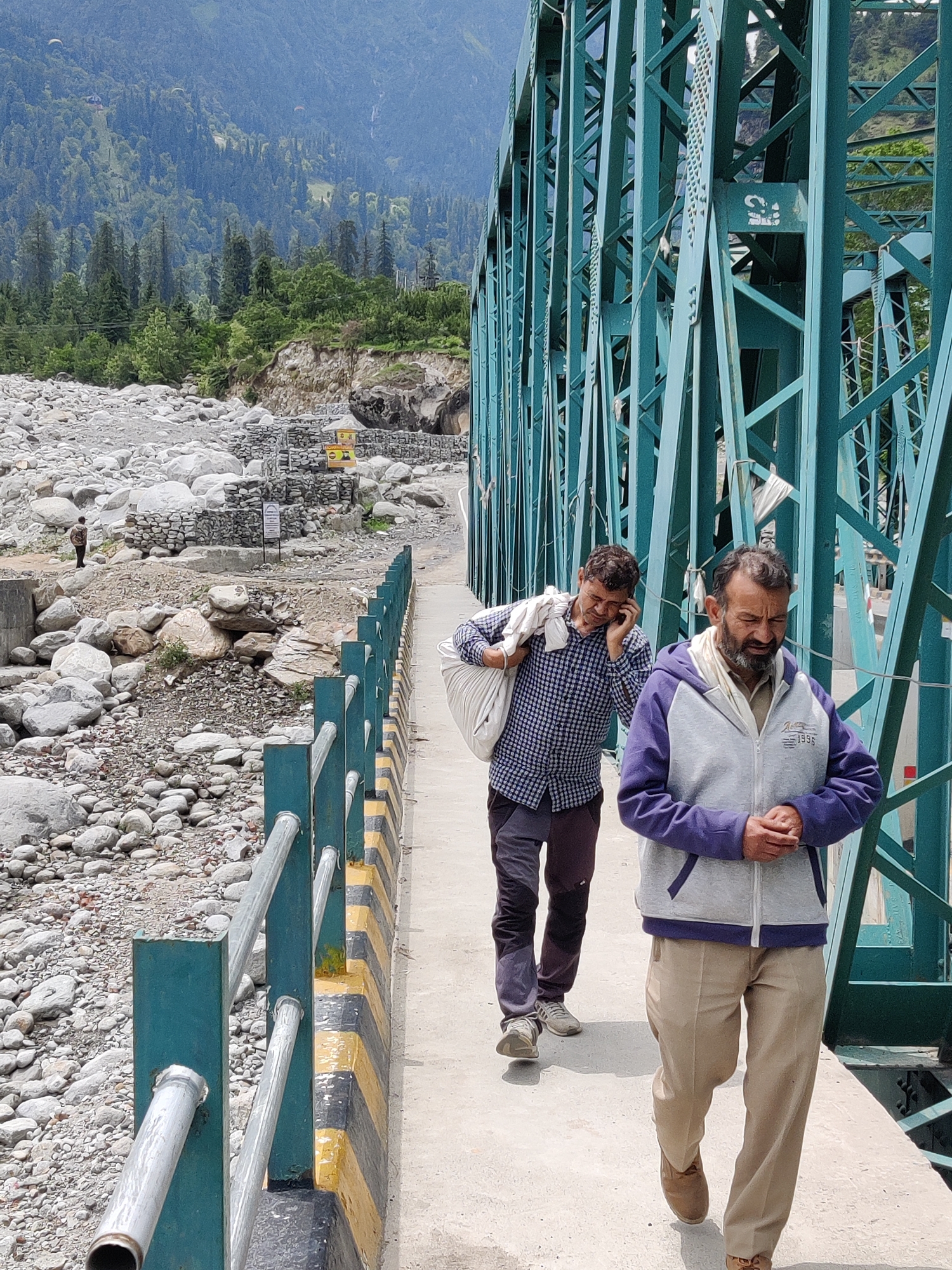 This is a full shot of a bridge in Palchan, India. Two men are walking across the bridge, which is painted teal. The man in the foreground is wearing a gray and purple hooded jacket and tan pants. He has his hands clasped in front of him. The man behind him is carrying a large white bag over his shoulder and is talking on a cell phone. He is wearing a blue and white checkered shirt and dark pants. The bridge spans over a rocky riverbed, and in the background, there are green, tree-covered mountains.