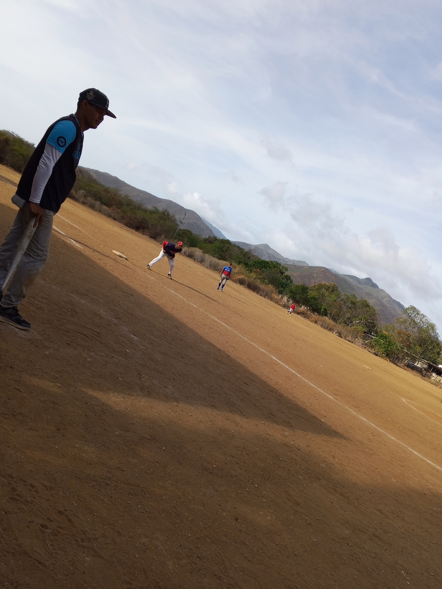 The image depicts a baseball game in progress on a dirt field, likely an amateur or informal setting. A man wearing a baseball cap, a black and blue top with long white sleeves, and gray pants stands in the foreground, holding a baseball bat. He is positioned near the first baseline. In the background, three players are visible on the field. One player in a red and white uniform is near first base, another player in a blue and red uniform is further out in the field, and a third person in red is more distant. The field is brown dirt with visible white baselines. Beyond the field, a mountain range is visible under a cloudy sky. The location is Unknown, Unknown.