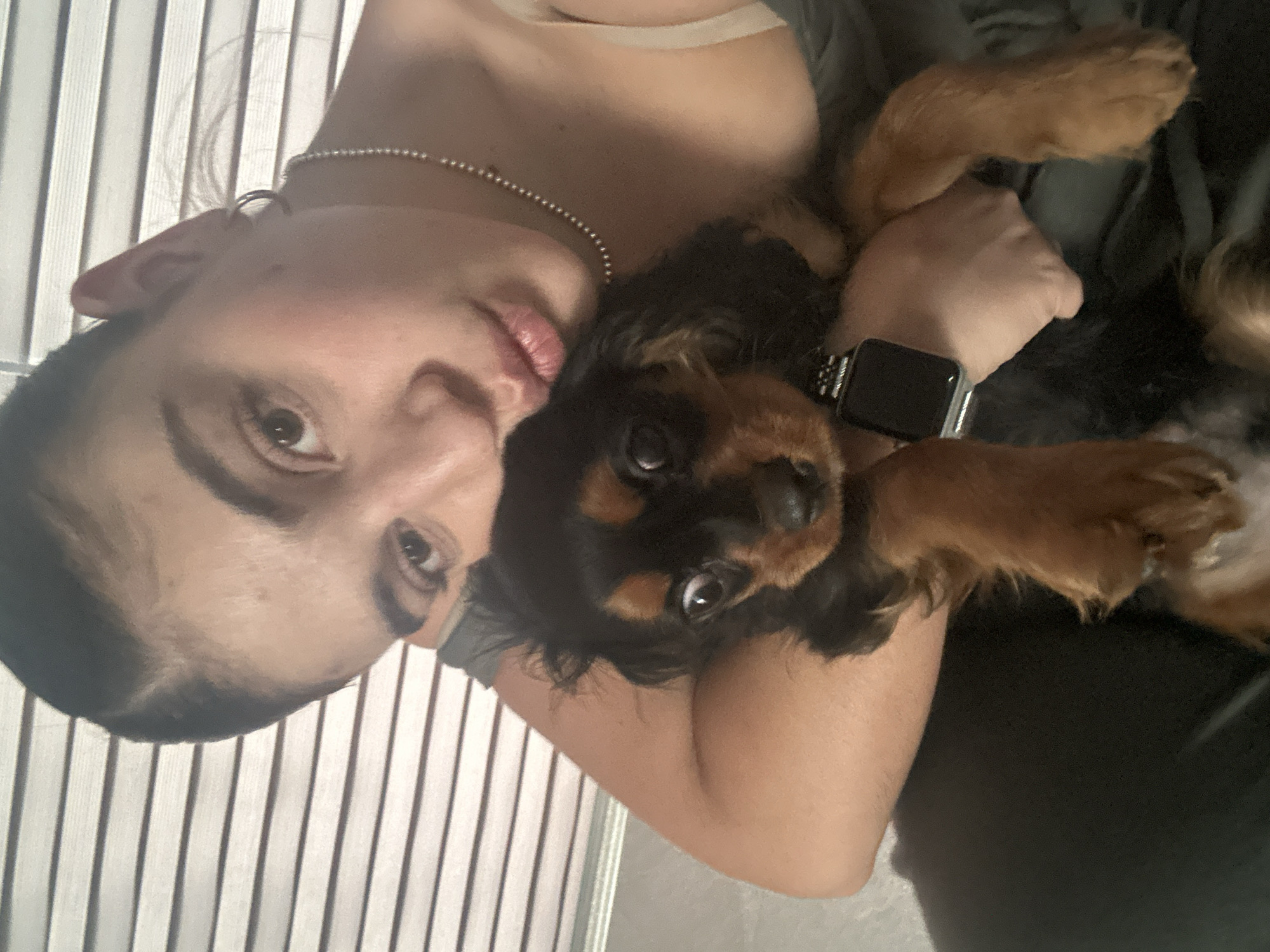A woman and a Cavalier King Charles Spaniel are in the frame. The woman is fair-skinned, has dark hair, and is wearing a silver chain necklace. She is looking directly at the camera. The dog, predominantly black and tan, is being held by the woman. A smartwatch is visible on the woman's wrist. Vertical blinds are in the background. The location, Puebla, Mexico, cannot be determined from the image.