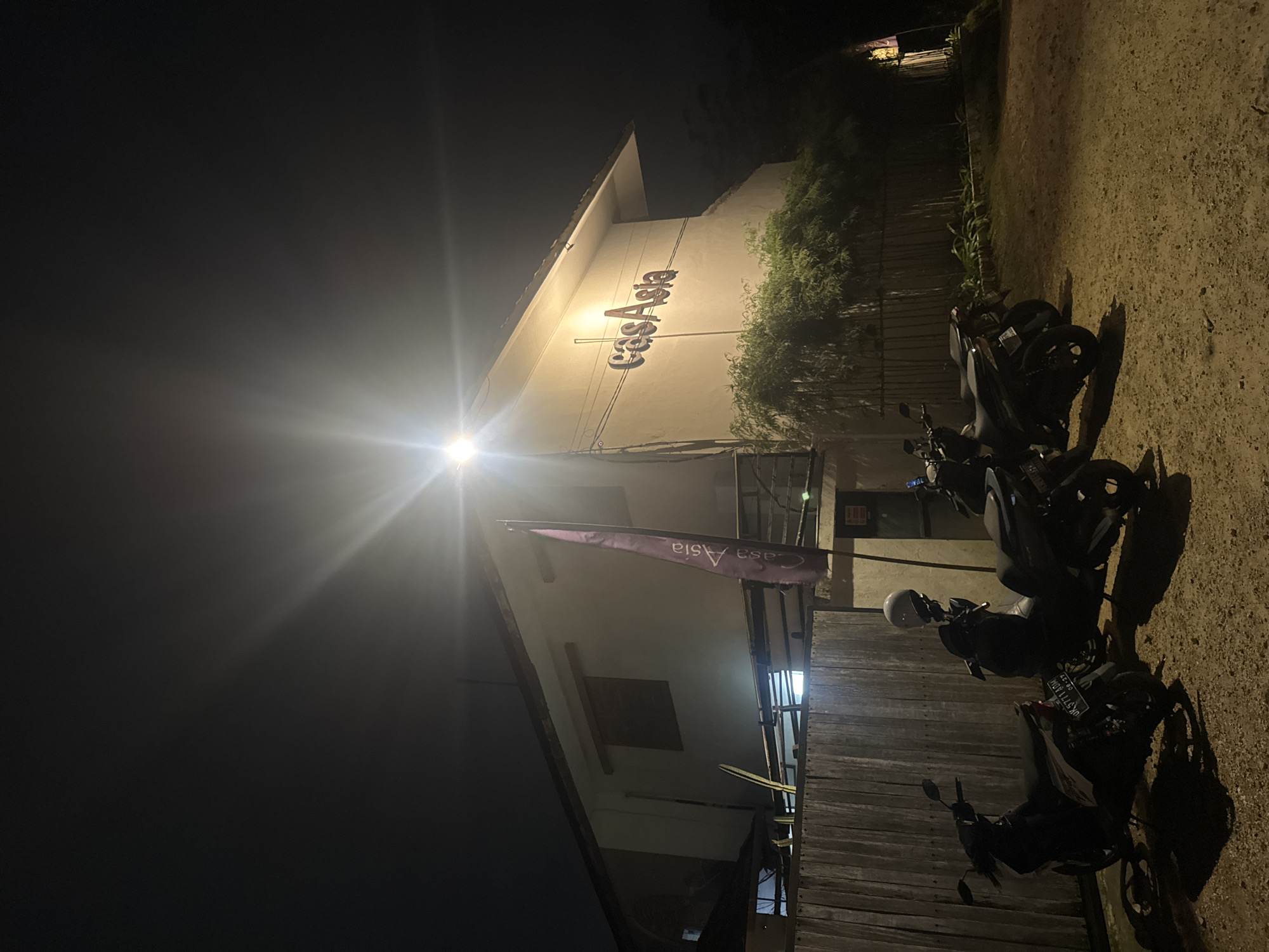 In the city of Kuta Selatan, Indonesia, at night, a well-lit building with the text "casaAsia" prominently displayed on its facade stands under a bright overhead light source. To the left of the building, a wooden fence runs horizontally across the foreground. Parked in front of the fence are three scooters, two of which are dark-colored and one is lighter colored with a white helmet on its seat. Behind the fence, lush green foliage and bamboo stalks are visible. A purple flag with white text is mounted on a pole to the right of the building. The ground appears to be dirt or gravel. The overall scene depicts a commercial establishment at night, likely a guesthouse or restaurant, with its entrance area illuminated.