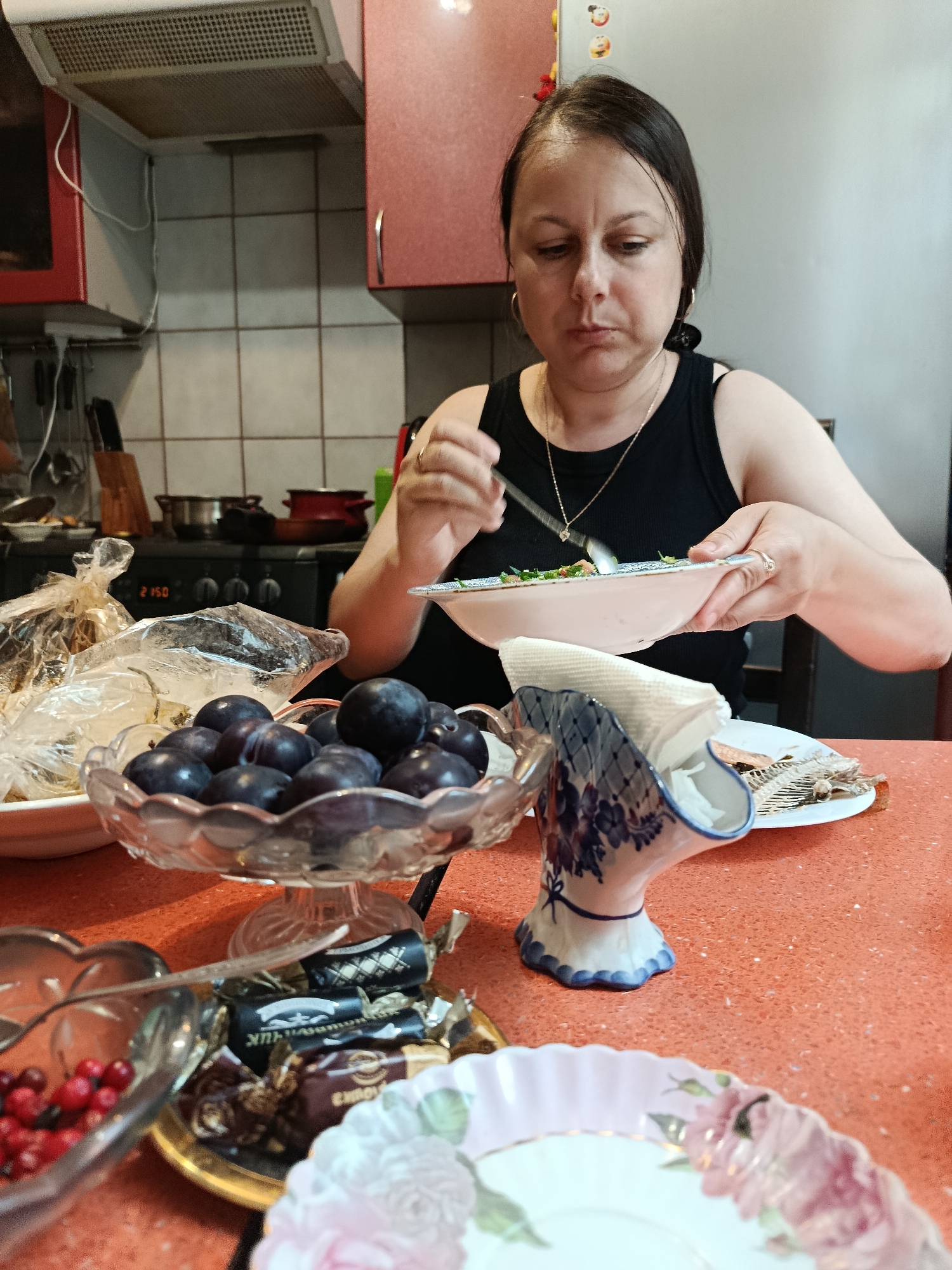 A middle-aged woman with dark hair is sitting at a table in a kitchen and eating from a plate. She is wearing a black tank top and a gold necklace. In front of her are bowls of plums and cranberries, as well as wrapped candies. Behind her, a stovetop and range hood are visible. The woman is holding a fork in her right hand and a plate in her left hand, and she appears to be in the process of eating. The kitchen appears to be a home kitchen, likely in a residential setting. The scene does not provide specific details to determine the location in Moscow, Russia.