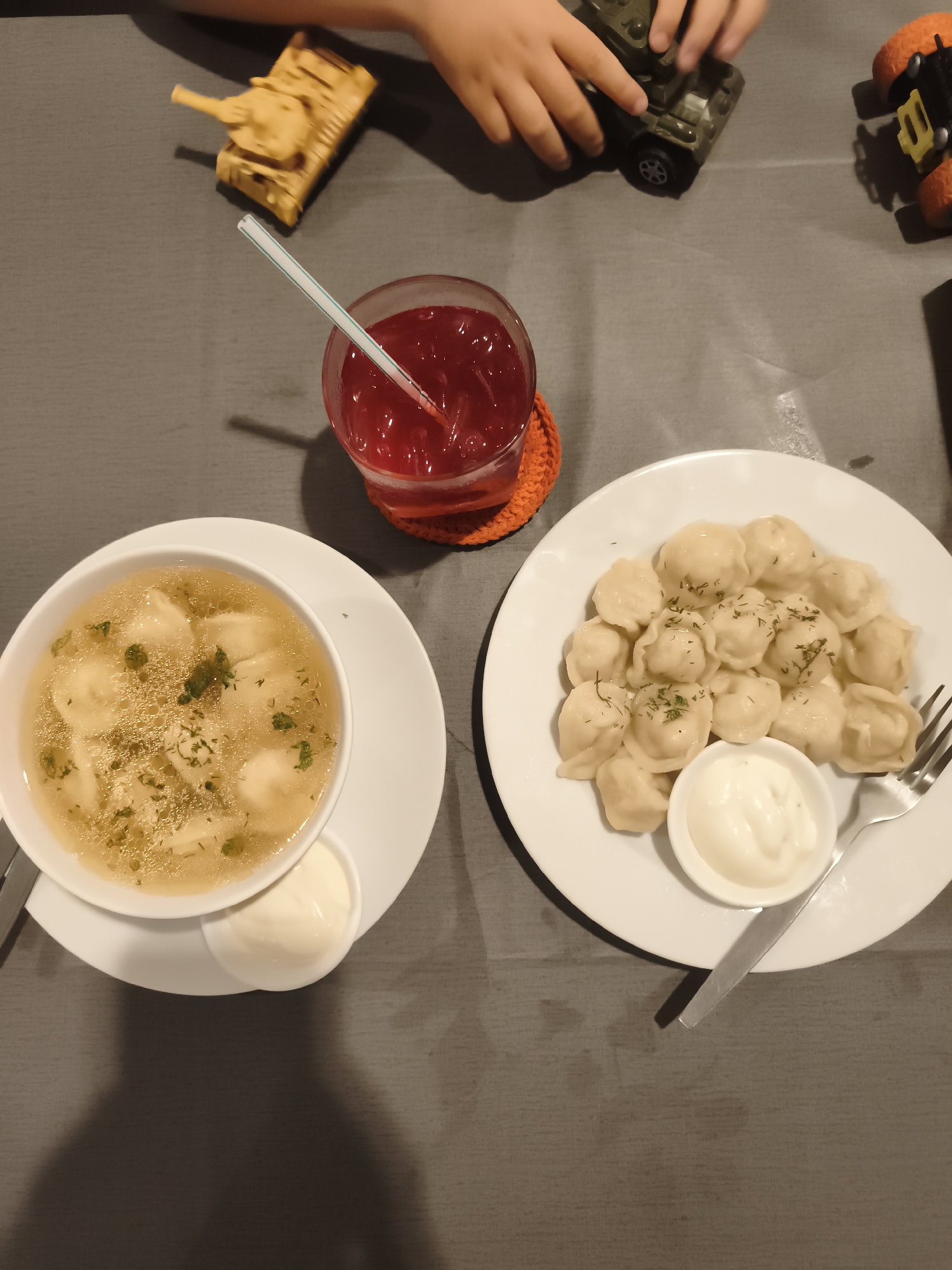 An overhead shot shows a meal on a gray tablecloth, with toys in the background. To the left, a white bowl contains broth with dumplings and green herbs, next to a small dish of sour cream. A red drink with a straw sits on an orange coaster. To the right, a white plate is piled high with dumplings, with a small dish of sour cream and a fork to the side. In the background, several toy vehicles are visible. The location is not specified in the image.