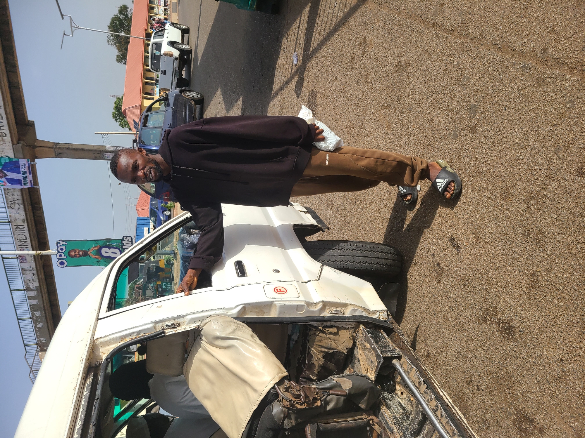 The image depicts a man standing next to a damaged white van in Gura Top, Nigeria. He is wearing a black hoodie, brown pants, and Adidas sandals, and has a bag in his hand. The van's passenger door and rear are severely damaged, exposing the interior. The setting appears to be a street with other vehicles parked in the background. A banner featuring a smiling man with an "OPay" logo is visible on a nearby bridge.