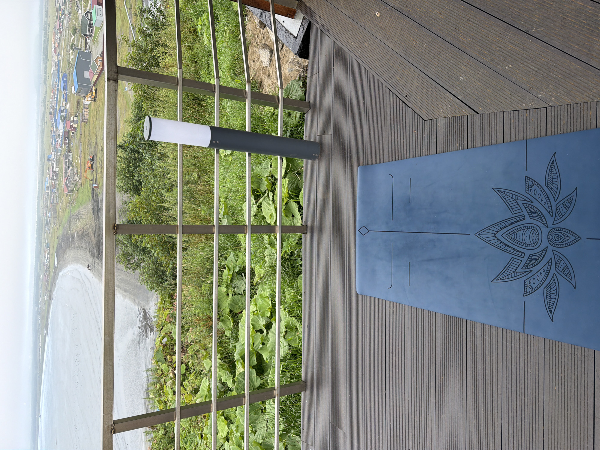 This image shows a blue yoga mat with a lotus flower design placed on a wooden deck. In the background, there is a metal railing overlooking a coastline. Beyond the railing, a town with various buildings is visible, situated near the sea. The sky is overcast. Based on the architecture and landscape, this location is likely Yuzhno-Kurilsk, Russia. There are no people visible in the image.