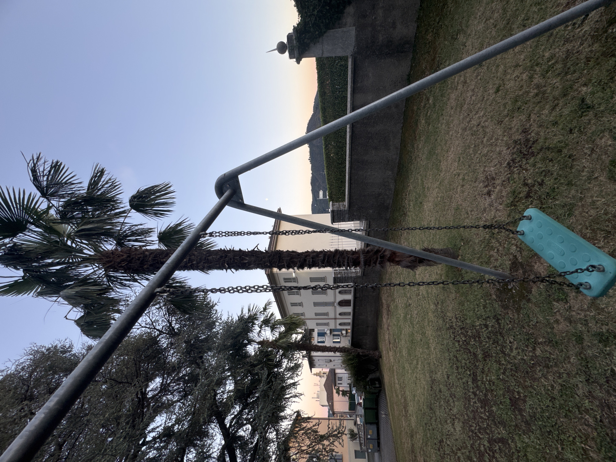 The image depicts a playground swing set with a teal seat hanging from metal chains, situated on a grassy area. In the background, there are several buildings, including a multi-story white building with arched windows and balconies, and other residential structures. Palm trees and other greenery are visible, as is a stone wall with a decorative finial. The sky is clear with a gradient of light blue to a pale orange near the horizon, suggesting either dawn or dusk. The overall scene appears to be in a residential area within the city of Balerna, Switzerland. There are no people visible in the image.
