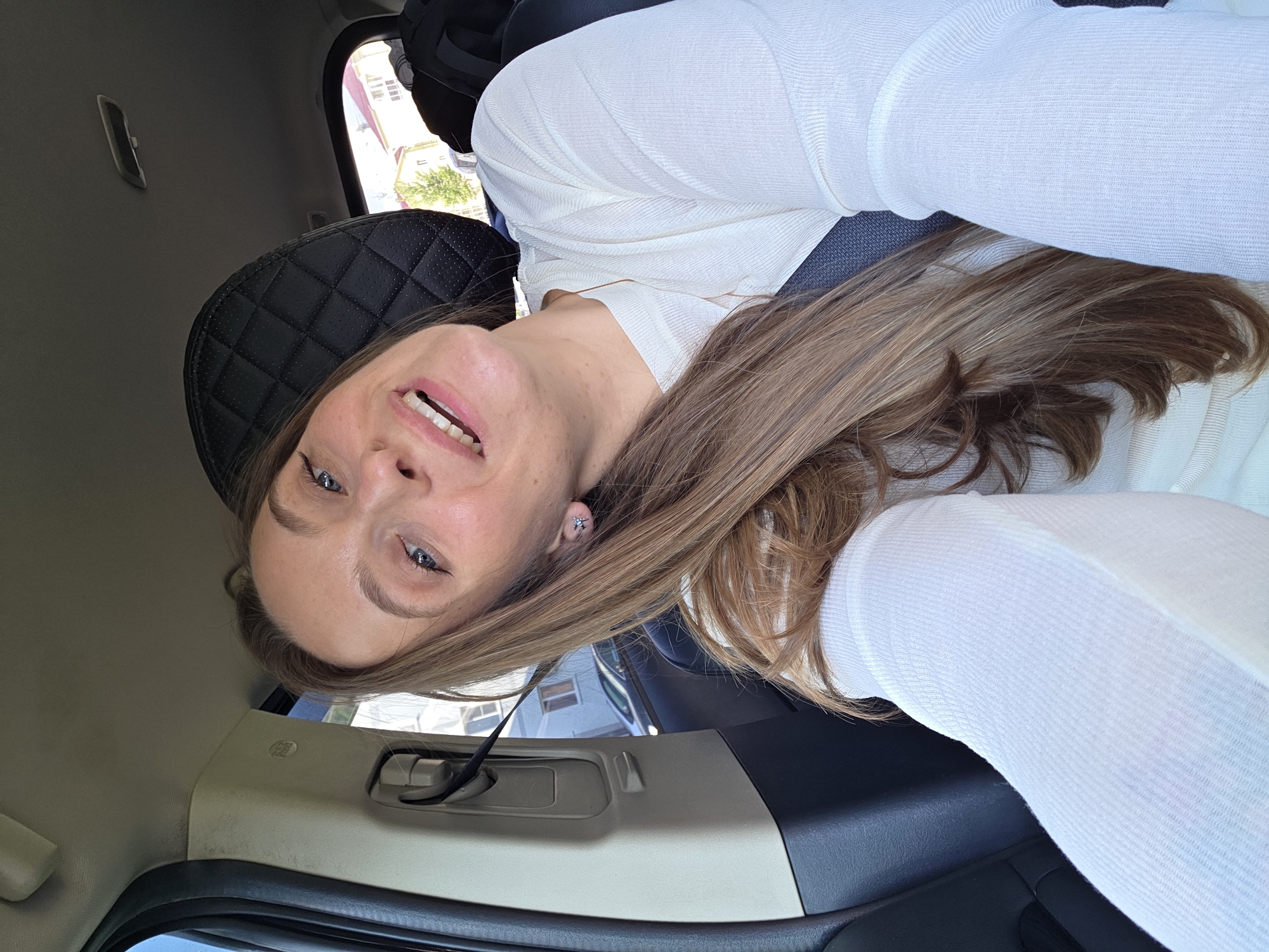 A young Caucasian woman with long, light brown hair and blue eyes smiles as she takes a selfie inside a vehicle. She is wearing a white, long-sleeved top and a seatbelt. The interior of the car is visible, including a black headrest and a grey interior panel. Outside the car window, buildings and trees can be seen, suggesting an urban or suburban setting. The lighting indicates it is daytime. The image does not provide specific details to confirm the location as Birobidzhan, Russia.