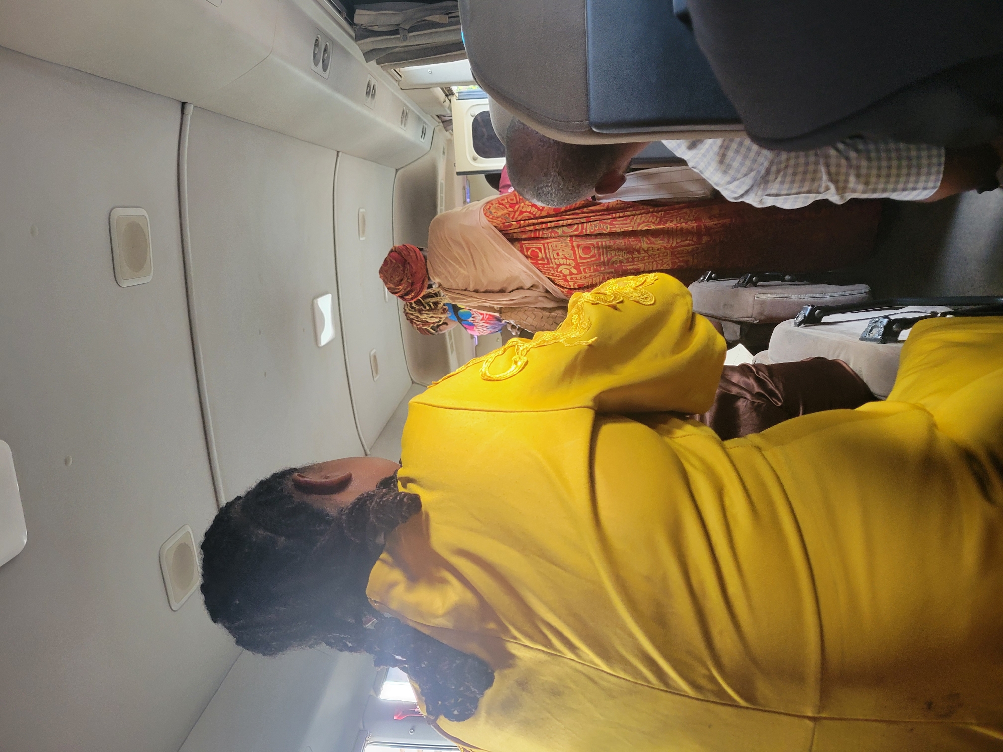 The image captures the interior of what appears to be an aircraft cabin, seen from a perspective that is slightly rotated, with the cabin walls running horizontally across the frame.
In the foreground, the back of a person wearing a bright, mustard-yellow garment is prominent. This individual has dark, curly hair. Moving towards the mid-ground, another person is seated with their back partially visible, wearing a light-colored, possibly beige or cream, head covering that resembles a turban, and a garment with red and gold patterns. Behind this person, the back of a man's head and the upper part of his light-colored, checkered shirt are visible as he sits.
The setting is clearly inside an airplane. Dark grey or black seats are visible in the immediate foreground, while seats further back appear to be upholstered in a lighter, possibly brown or beige, material. The cabin walls are a light grey or white, featuring small, rectangular panels that could be speakers or call buttons. Overhead compartments are visible along the ceiling, and a narrow aisle can be inferred between the rows of seats. The overall impression is that of a passenger compartment in a smaller aircraft, possibly a regional jet or turboprop, given the relatively compact nature of the interior.
The activity taking place is that of passengers seated in an aircraft, likely either awaiting departure, during a flight, or after landing. The time of day appears to be daylight, as indicated by the general illumination of the cabin, although no external windows showing the outside are clearly depicted. There is no visible text in the image. The image is taken within the context of Abuja, Nigeria.