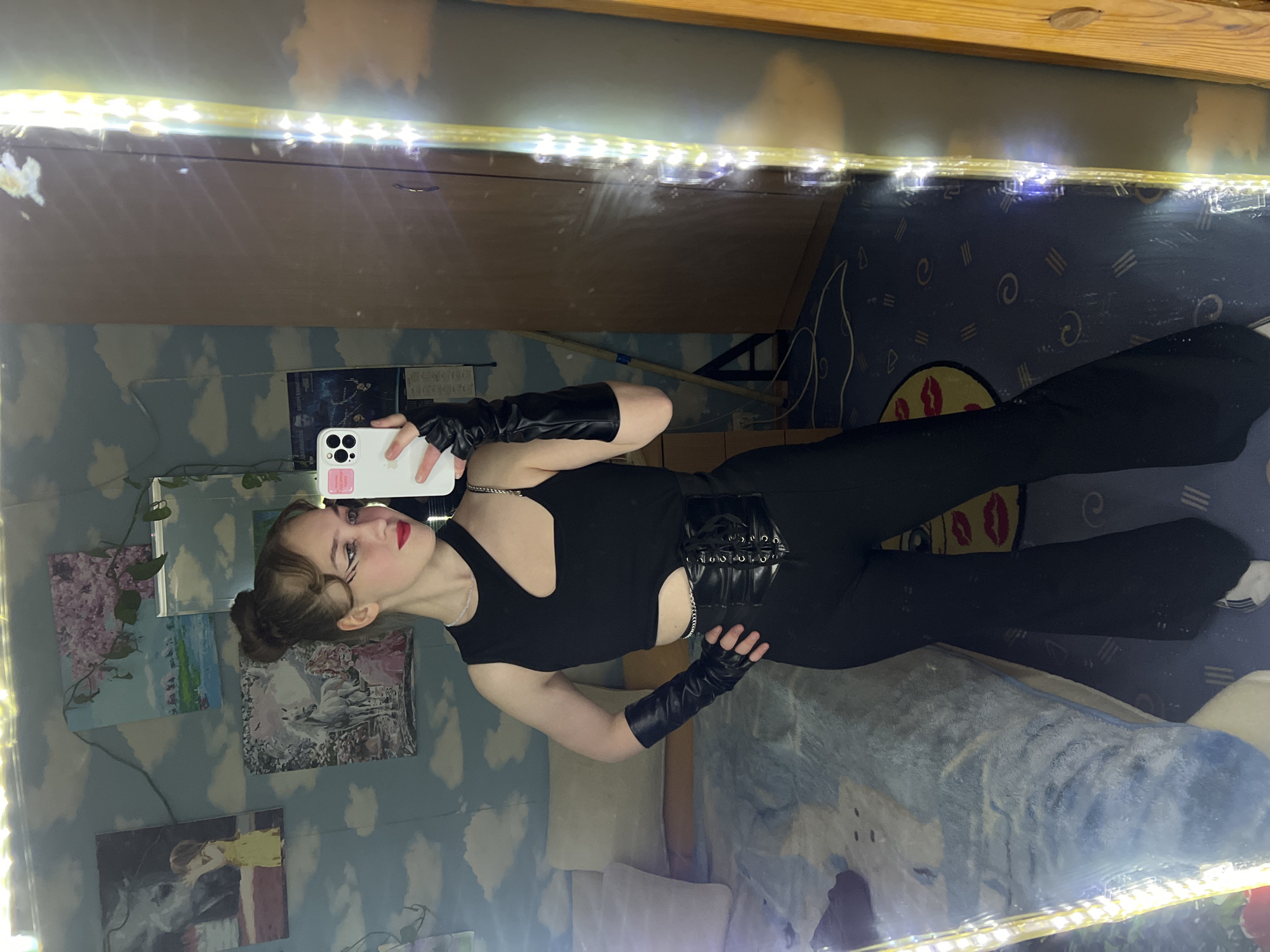 A woman in a black outfit, including gloves and a corset-style belt, is taking a selfie in a mirror. The mirror is surrounded by LED lights. The room has a blue wall with cloud patterns and paintings, and a bed with blue and white bedding is visible. The exact location is unknown.