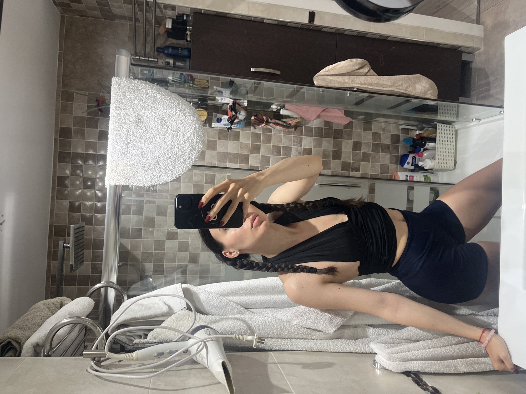 A young woman with dark hair in two braids is taking a mirror selfie in a bathroom. She is wearing a black halter top and dark blue shorts. The bathroom has tiled walls, a shower with a glass door, and a cabinet filled with toiletries. There is a hairdryer hanging on the wall and several towels are visible. The woman's left arm has a pink bracelet on her wrist. The image does not provide any information that would suggest the location is in Palma, Spain.