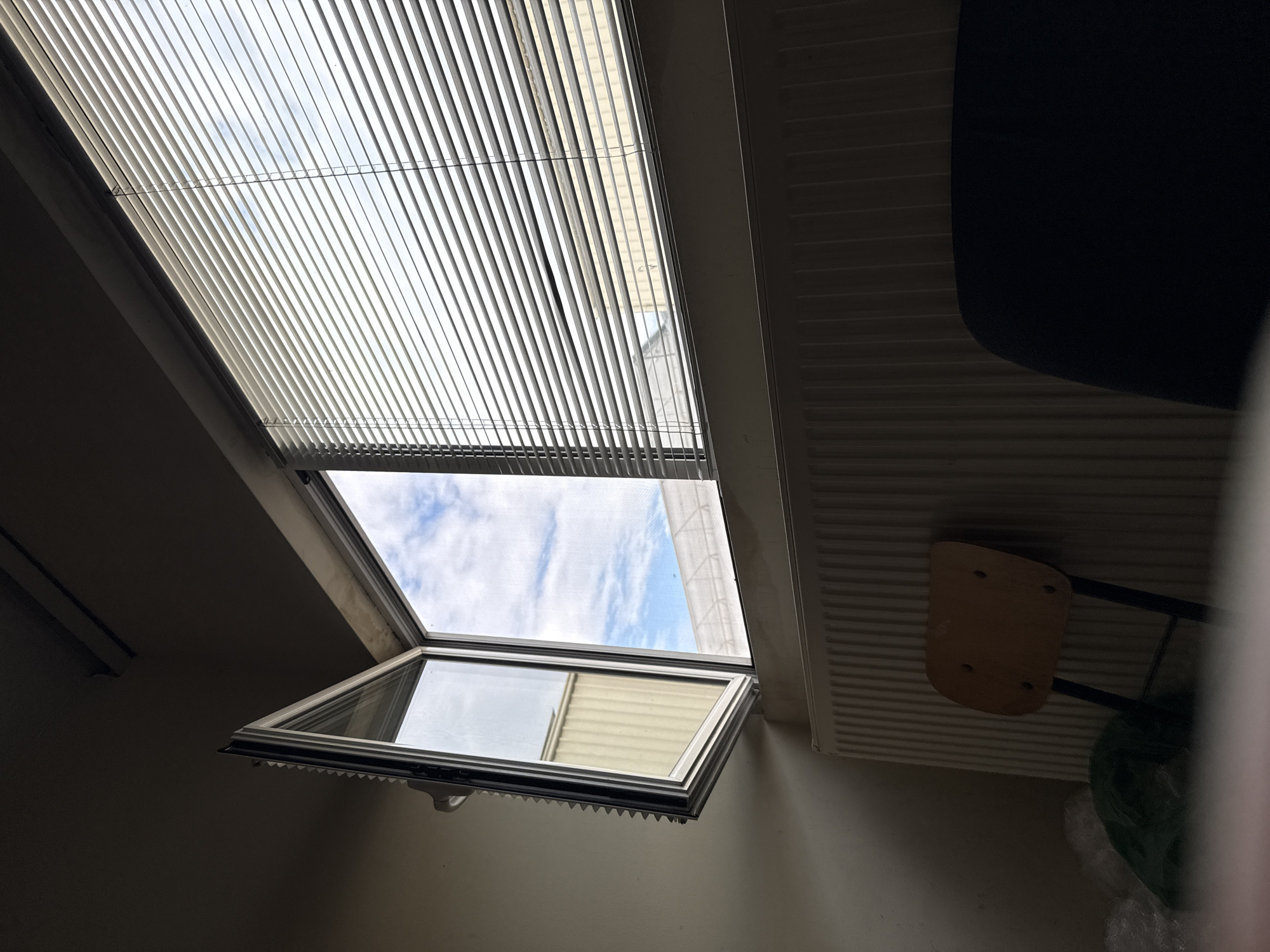 The image captures an interior view of a room in Stara Zagora, Bulgaria, looking upwards towards a roof window (skylight). The upper section of the skylight is covered by white, horizontal Venetian blinds, which are partially drawn, allowing diffused daylight to filter into the room. A thin wire runs across the blinds, likely part of the operating mechanism.
Below the blinds, the lower pane of the skylight is open, tilted downwards into the room. Through this open window, a bright, cloudy blue sky is clearly visible. Reflections in the glass of the open pane show parts of an exterior building structure, possibly siding or a light-colored wall.
To the right of the window, a white, horizontally ribbed radiator is mounted on a wall. Below and slightly in front of the radiator, a small, light-colored wooden chair seat with dark metal legs is visible, appearing to be turned on its side or leaning. In the upper right corner of the frame, a dark, indistinct object, possibly a part of furniture, is partially visible.
The scene depicts a daytime setting with natural light entering the room through the window and blinds, creating a contrast with the relatively darker interior. No people or visible text are present in the image.