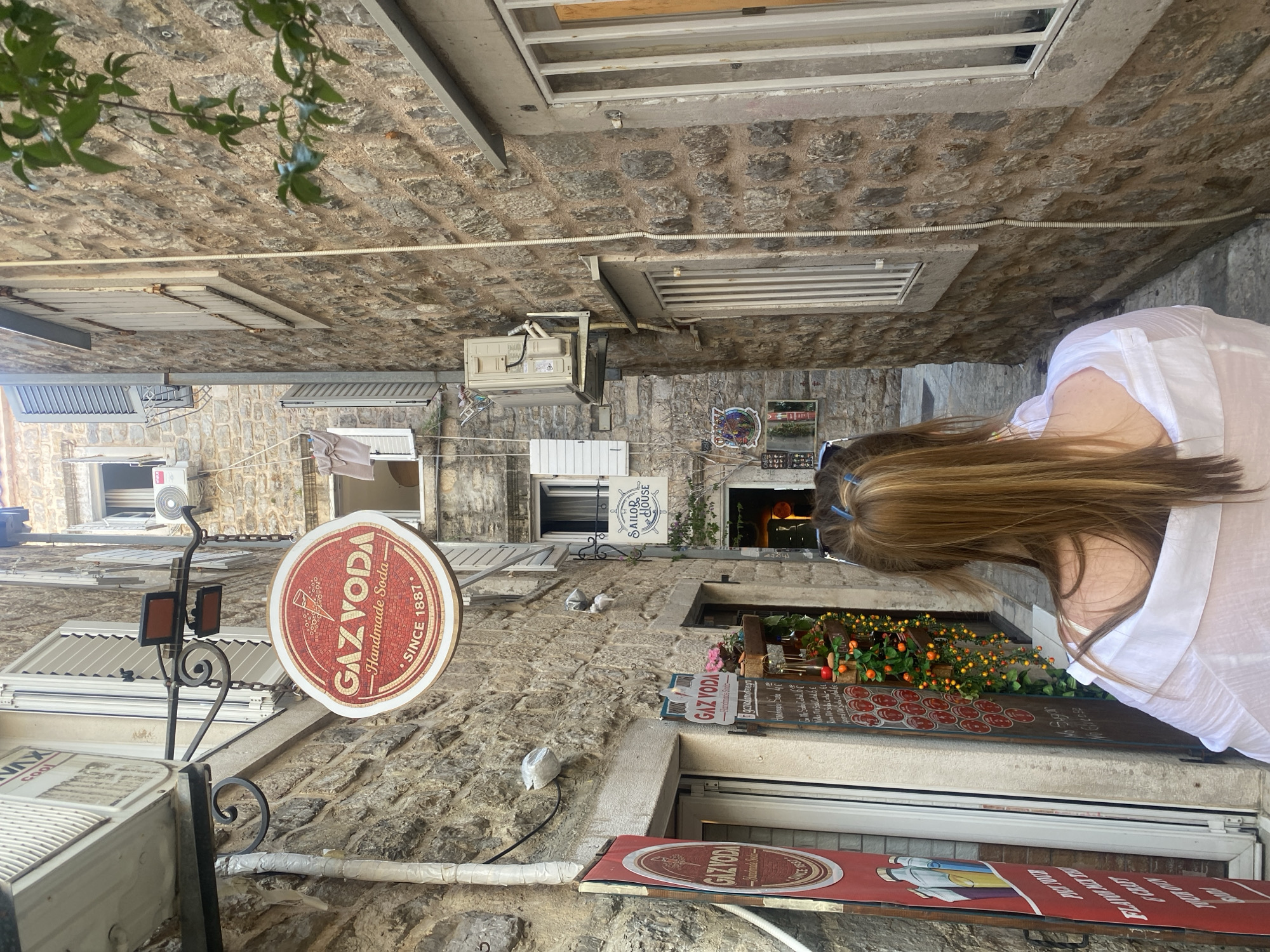 An eye-level, candid daytime shot depicts a narrow cobblestone street in Budva, Montenegro. Weathered stone buildings line both sides, interspersed with windows covered by white louvers.
On the left side of the street, a black wrought-iron sign holder supports a circular advertisement sign, displaying the brand name "GAZYODA" in red lettering around a vintage soda glass graphic, alongside the words "Handmade Soda- SINCE 1887". Below this, smaller signage is visible, accompanied by an air conditioning unit and other fixtures attached to the stone wall.
To the right, an indistinct woman with long brown hair, wearing a white top, is partially in the frame. A storefront is partially visible, decorated with floral arrangements and a menu board. The words "GAZ VODA" appear on additional signage.
The perspective draws the eye down the street, revealing a succession of buildings with similar architectural characteristics, with a few plants overhead. A white wire is visible near the top of the frame, traversing the street between the buildings.