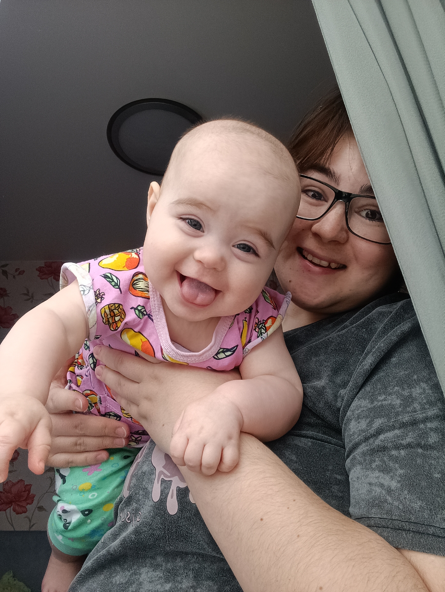 A woman holds a baby in her arms. The baby wears a pink top with fruit patterns and green patterned leggings, sticking its tongue out and smiling. The woman wears glasses and a grey t-shirt, also smiling. Part of a grey curtain is visible on the right side of the image. A ceiling with a light fixture is visible above the woman and the baby. Wallpaper with a floral pattern is visible in the background. This photograph was taken in an interior location, and due to the specified location in the prompt it is likely Noviy, Russia.