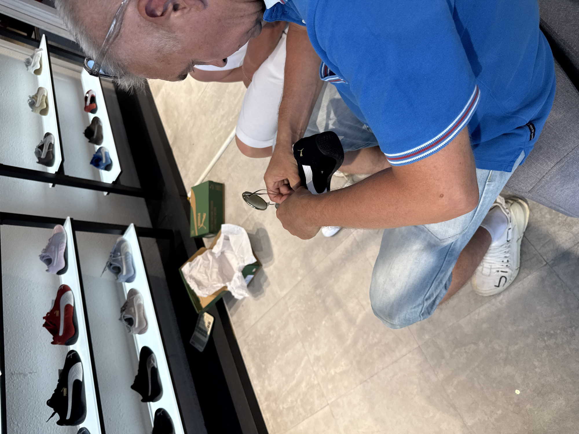 The image depicts a man sitting on the floor in a shoe store, examining a black and white Puma sneaker. He is wearing a blue polo shirt with red and white trim on the collar and sleeves, and light blue denim jeans. The man has gray hair and is wearing glasses. Beside him, a portion of another person wearing a white shirt is visible. In the background, shelves display various Puma sneakers in different colors. A green shoe box and crumpled white tissue paper are on the floor in front of the shelves. The floor is tiled with a light gray, patterned material. The scene appears to be inside a retail environment.