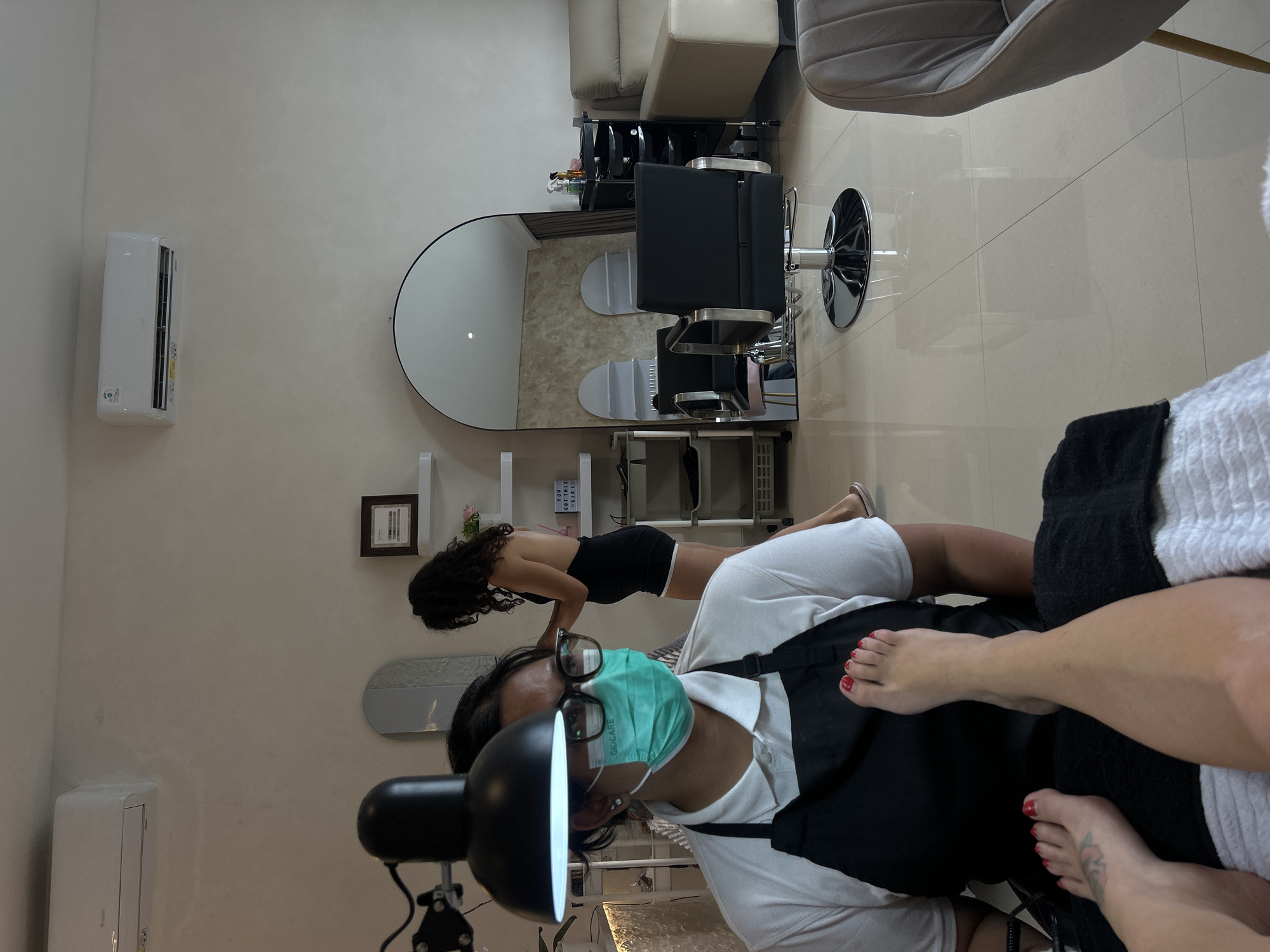 The image depicts a pedicure service at a beauty salon in North Kuta, Indonesia. A person wearing a white shirt, black apron, glasses, and a turquoise face mask is seated, with their feet extended in the foreground, one of which has red nail polish. Another person, a woman with dark curly hair wearing a black outfit, is standing in the background, seemingly attending to hair. The salon environment is visible, including an air conditioning unit, a large arched mirror reflecting chairs and shelving, a trolley with beauty products, and seating furniture. The flooring appears to be tiled.