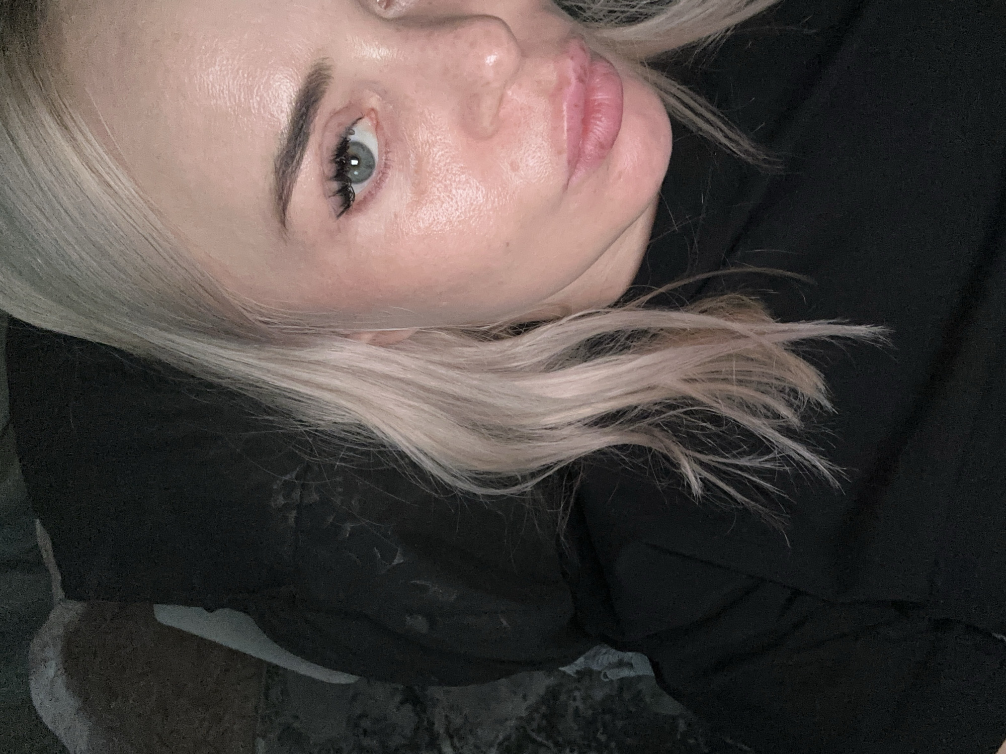 A close-up, selfie-style shot shows a woman with blonde hair and light skin. She is wearing a black shirt with a high neckline. Her eyes are open, and she is looking directly at the camera. Her lips are slightly parted. The background appears to be dark and textured, possibly a couch or chair. The lighting is soft and appears to be coming from the front. The image is focused on the woman's face and upper chest.