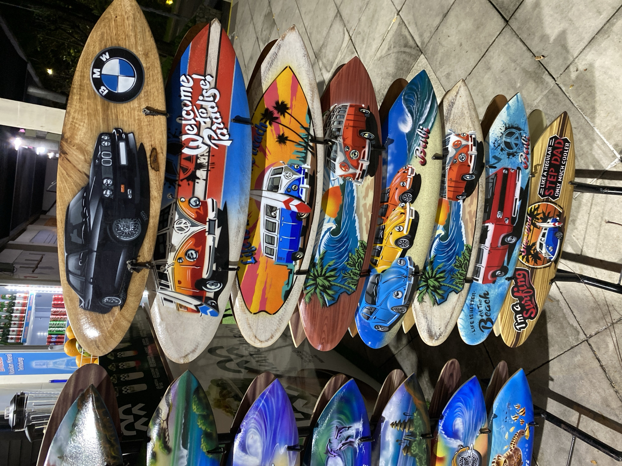 The image shows a display of surfboards with various artistic designs, predominantly featuring vintage Volkswagen vans, cars, and surf-themed graphics. The surfboards are arranged vertically on stands. In the background, a refrigerator stocked with drinks and some orange fruits are visible, suggesting a commercial setting. The overall scene appears to be an outdoor stall or shop, possibly in a tourist area given the surf theme. The lighting suggests it might be evening or nighttime. The ground is paved with light-colored tiles. Some of the designs on the surfboards include the text "Welcome to Paradise!", "Bali," "Life is Better At The Beach," and "Like A Regular Step Dad Only Much Cooler." One surfboard features a BMW logo and a black BMW car.