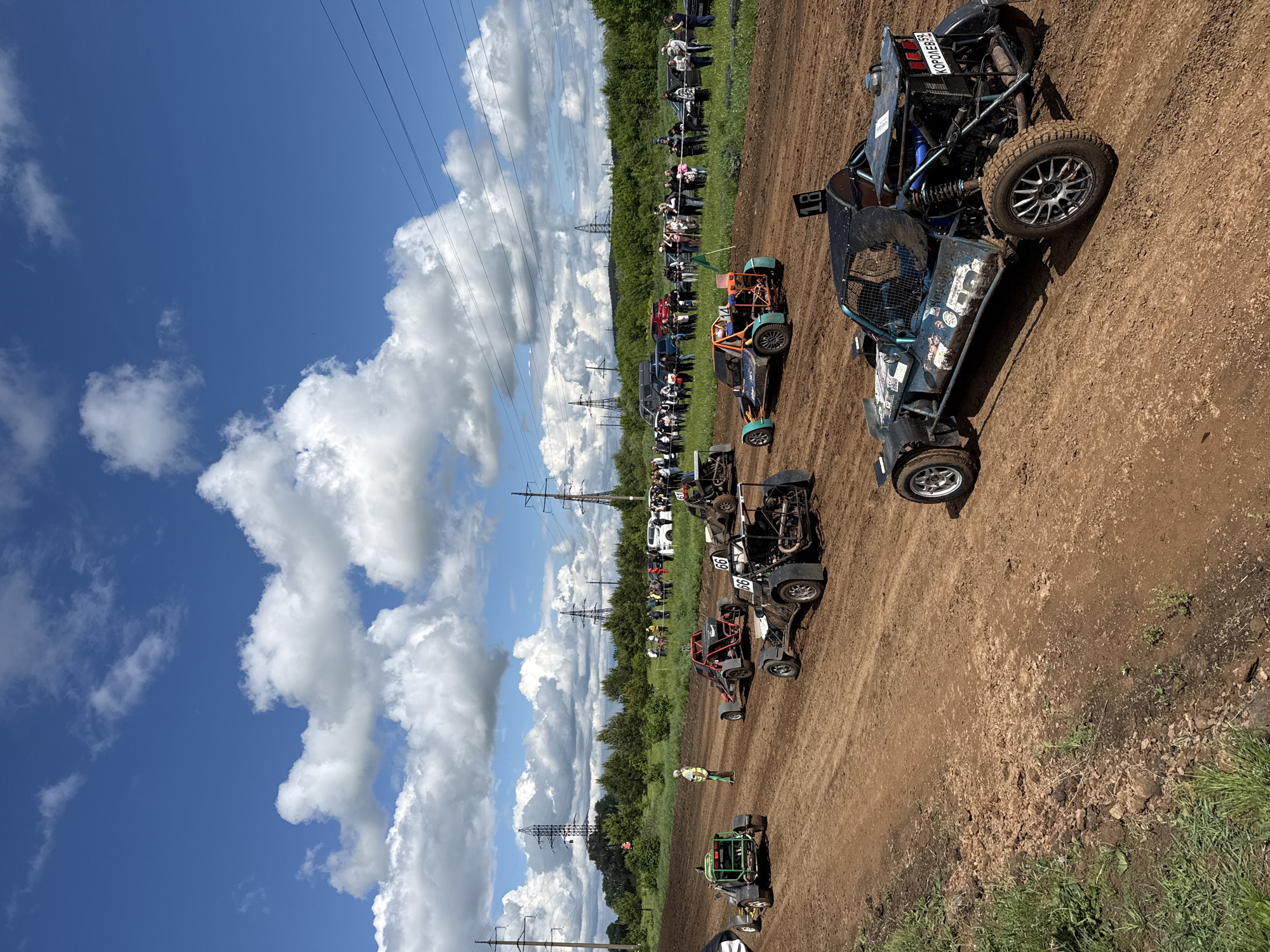 The image depicts a dirt track racing event. A line of racing buggies are positioned on a dirt track, ready to race. Spectators line the perimeter of the track, viewing the event. Power lines run along the side of the track. The location is in the city of Votkinsk, Russia.
